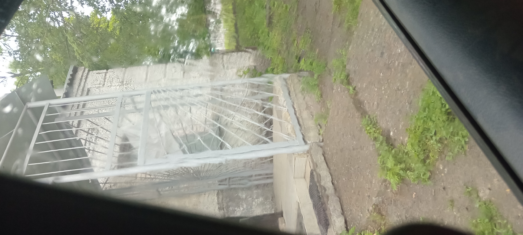 The image shows a view from inside a vehicle, looking out at a slightly blurry outdoor scene. The dominant objects are a light-colored, wrought-iron gate and a textured, light-colored wall behind it, possibly part of a building. The gate is elaborate with a decorative fan-like pattern at the bottom and horizontal bars above. Beyond the wall, there is lush green foliage, suggesting trees or bushes, and an area of unpaved ground with patches of green vegetation. The ground appears to be dirt and possibly gravel, with some areas looking damp, hinting at recent rain.
The scene is set in an outdoor environment, possibly a residential area or the grounds of a facility. The lighting suggests it is daytime, but the exact time is difficult to ascertain due to the blurriness and overcast appearance. The weather could be rainy or recently rained upon, given the damp ground. There are no people visible in the frame. The perspective is from a low angle, looking outwards and slightly upwards. The image is oriented with the top of the scene towards the left, implying the photograph may have been taken with the camera held at an angle or the vehicle was parked on a slope. There is no visible text in the image.