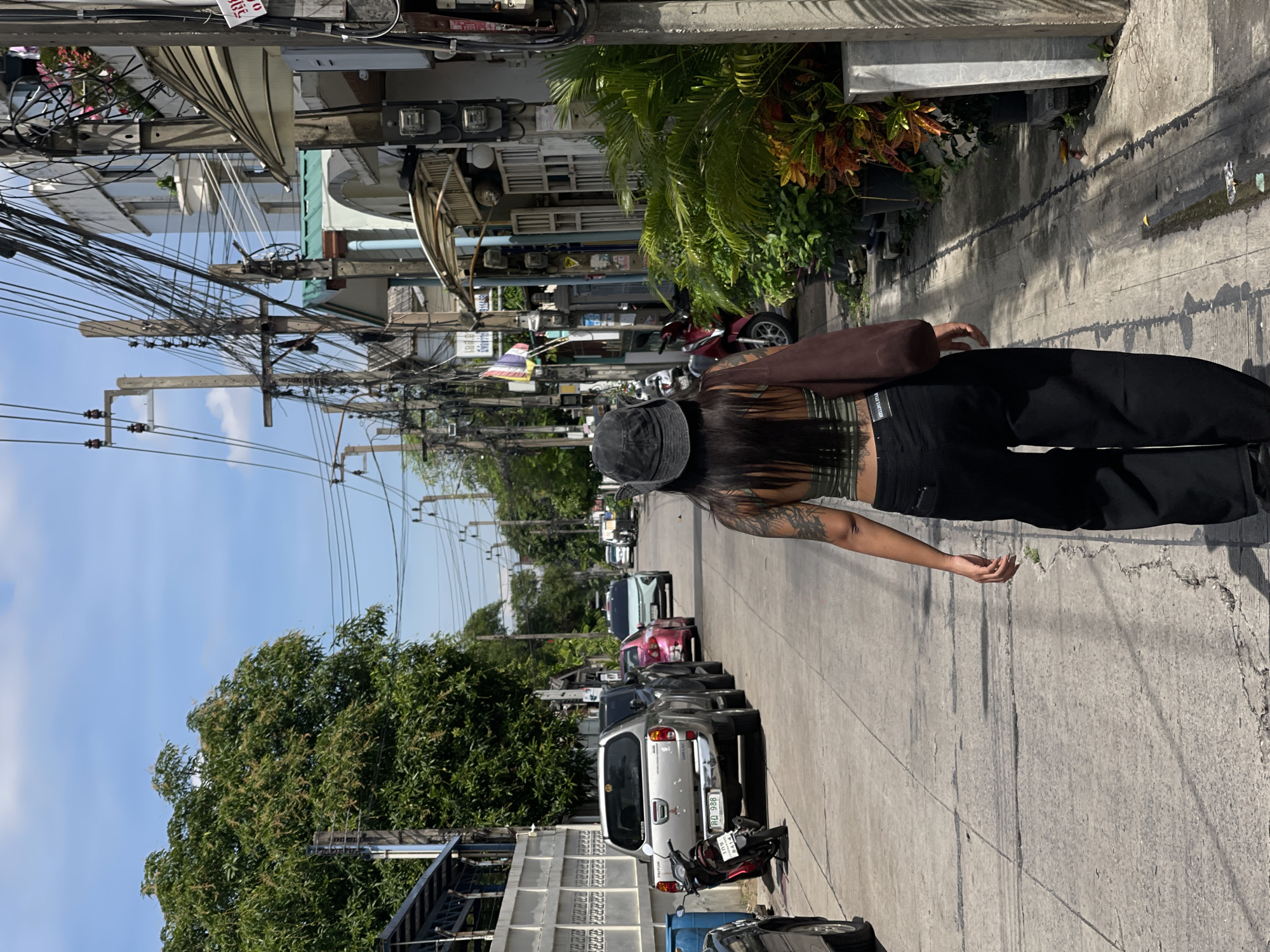 A person with long dark hair, wearing a dark bucket hat, a green and brown striped halter top, and black wide-leg pants walks away from the camera down a narrow city street in Bangkok, Thailand. The person has tattoos on their left arm and back. They are carrying a brown bag over their right shoulder. The street is paved with concrete and has visible cracks and stains. Several vehicles are parked along the side of the street, including a silver pickup truck, a red motorcycle, a pink sedan, and a light blue SUV. Buildings line both sides of the street, with some appearing to be shops or residences. Overhead, a complex network of electrical wires and poles crisscrosses the sky, typical of Bangkok's urban infrastructure. Lush green trees and potted plants add touches of nature to the otherwise urban landscape. The sky is blue with scattered white clouds. The scene is illuminated by bright sunlight, casting shadows on the street.