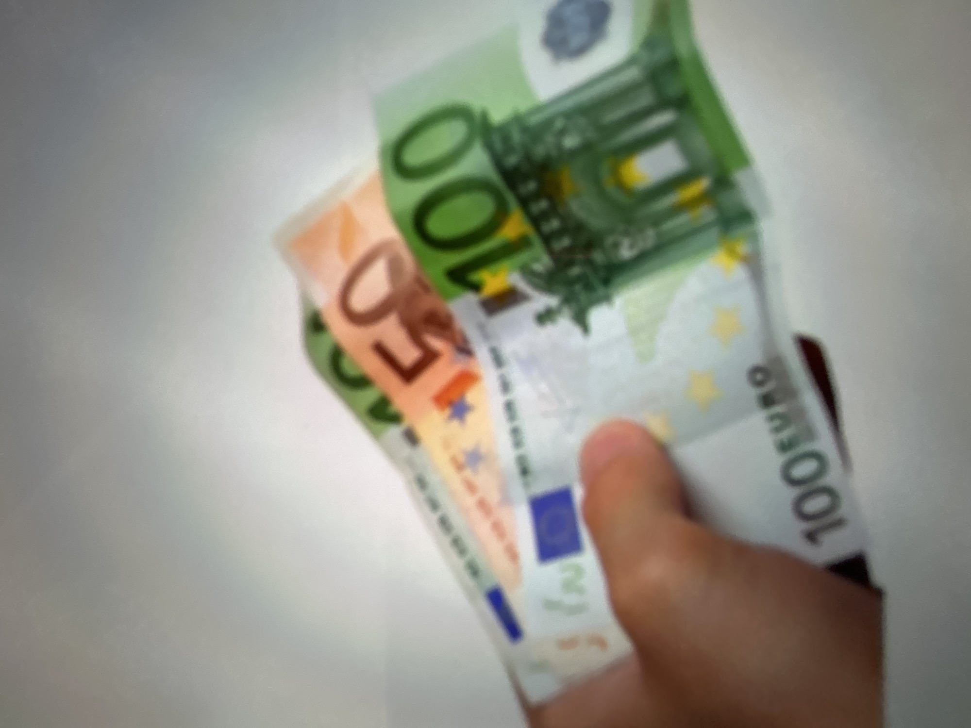 The media file displays a close-up, blurry image of a hand holding several euro banknotes. The hand appears to be lightly gripping the bills, which are fanned out. The visible banknotes are a 100 euro note, a 50 euro note, and what appears to be a 20 euro note. The 100 euro note is predominantly green, while the 50 euro note is orange. The background is out of focus, showing a light, neutral surface, possibly a table or wall. There are no clear indicators of time of day, weather, or specific location beyond the general context of euro banknotes. The image captures a simple act of holding money, with no discernible emotional cues or specific event taking place. Visible text on the banknotes includes "100 EURO", "50 EURO", and what appears to be "20 EURO". The euro symbol (€) is also visible on some of the notes. The setting is a generic indoor environment.