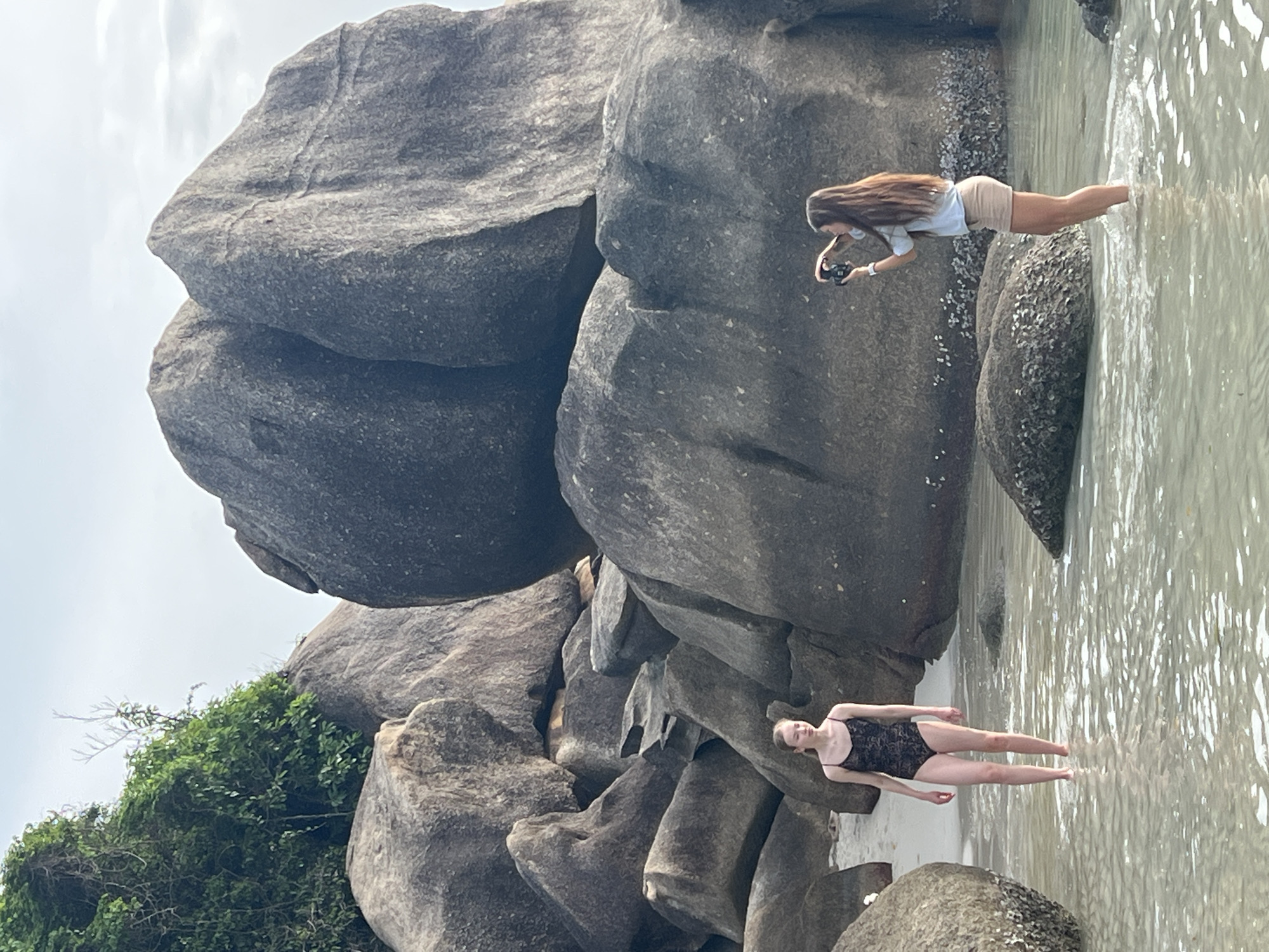 Two young women are standing in shallow, clear water on a beach with large, smooth, gray boulders. On the left, a woman in a black, lace-patterned swimsuit stands facing slightly to the right, looking forward. On the right, a woman with long brown hair, wearing a light blue long-sleeved shirt and tan shorts, is taking a photograph of the woman on the left with a black camera. The background is dominated by the massive boulders, with some green foliage visible on the upper left. The sky is overcast. This scene is consistent with a location in Baan Chaweng Noi, Thailand, known for its picturesque beaches and rock formations.