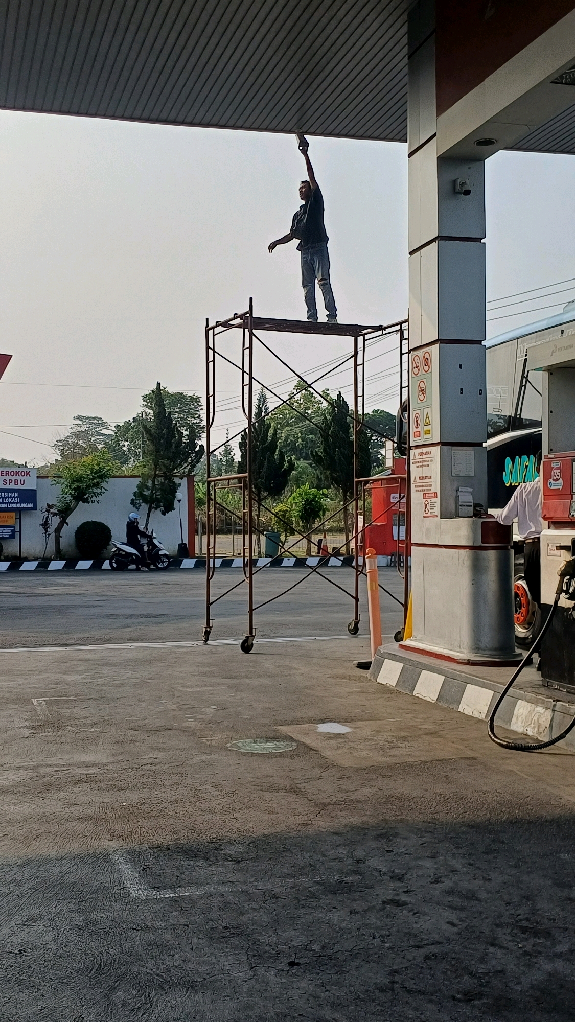 In Kranggan, Indonesia, a man is standing on a metal scaffolding at a gas station, holding a spray can and seemingly painting the underside of the station's roof. He is wearing a dark t-shirt and light-colored, ripped jeans. The scaffolding is positioned on the asphalt ground. In the background, a person on a motorcycle is visible on the road. A gas pump station with signage and a large vehicle are also present. The scene is illuminated by natural daylight, casting shadows on the ground.