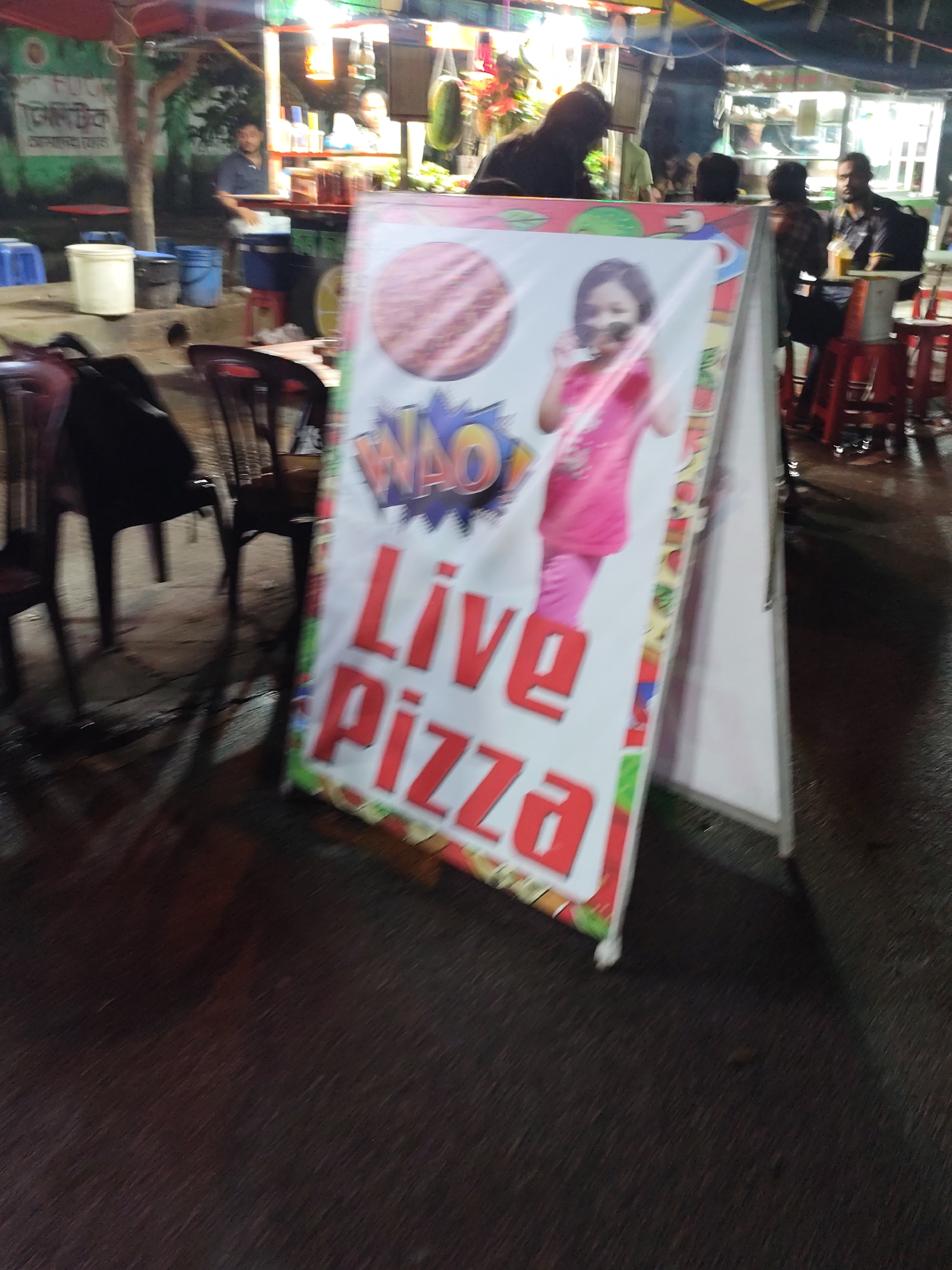 The image depicts a street-level view of a food stall in Dhaka, Bangladesh, at night. A prominent A-frame sign, a common sight in such markets, displays the words "Live Pizza" in large red lettering against a white background. Above the text, a cartoonish graphic appears, possibly related to the food being advertised. The sign also features a depiction of a young girl in a pink top.
In the background, the food stall itself is illuminated by bright lights, suggesting an active business. Several people are visible, some working behind the counter and others as patrons. One person is seated at a table with a drink, while others are standing. The environment appears to be an open-air setting, common in Dhaka's bustling street markets, with the ground being a dark, wet surface, indicative of recent rain or spillage. Chairs and tables are set up for customers. The overall impression is of a lively street food scene.