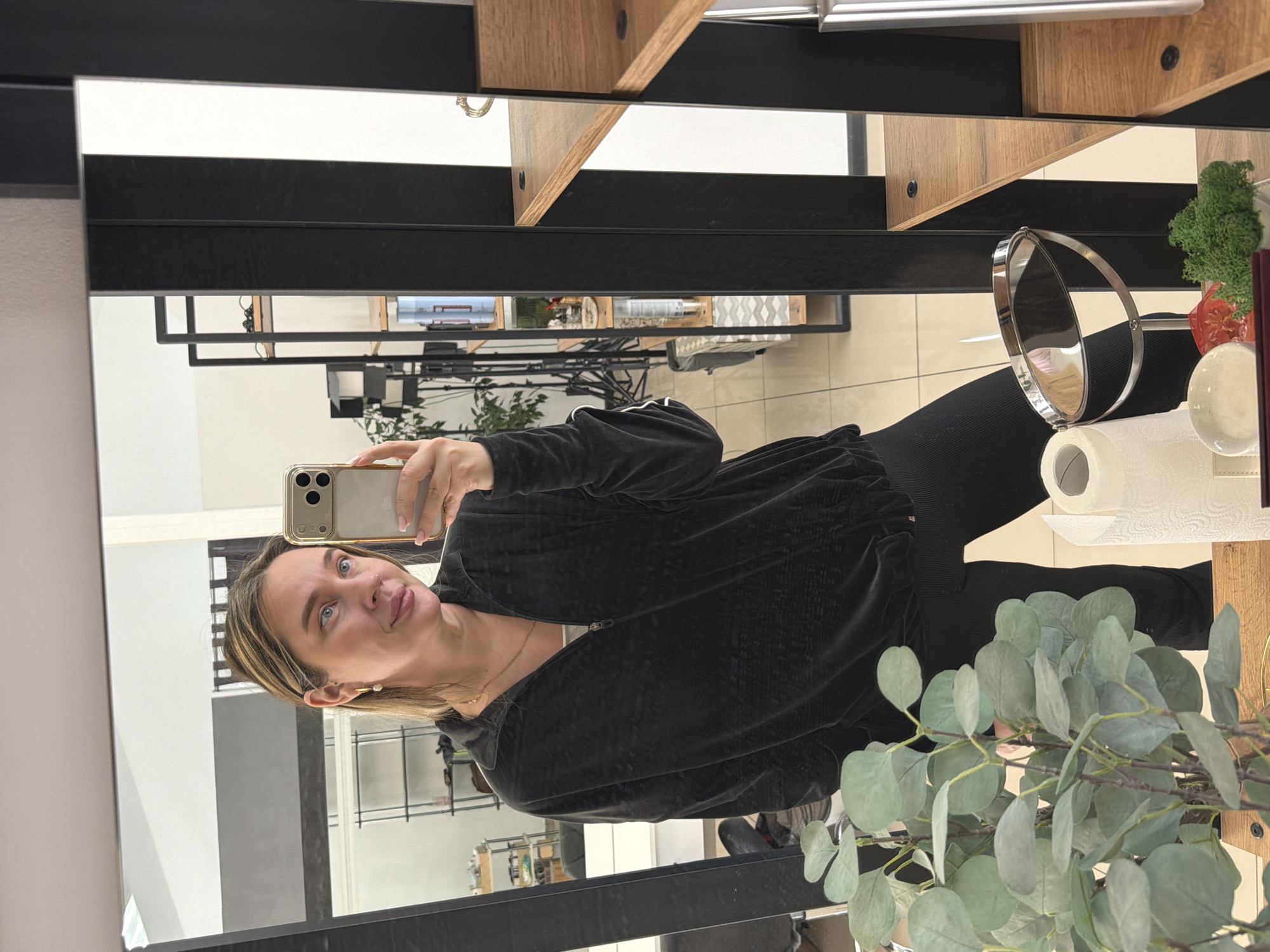 A woman is taking a selfie in a mirror. She is wearing a black jacket and black pants. She has blonde hair and blue eyes. The mirror is in a room with shelves and plants. There is a small mirror with a roll of toilet paper next to it. The location is likely a business, potentially a salon or retail space, but the specific location in Chaikovsky, Russia cannot be determined from the image. The woman is holding a gold-colored smartphone.
