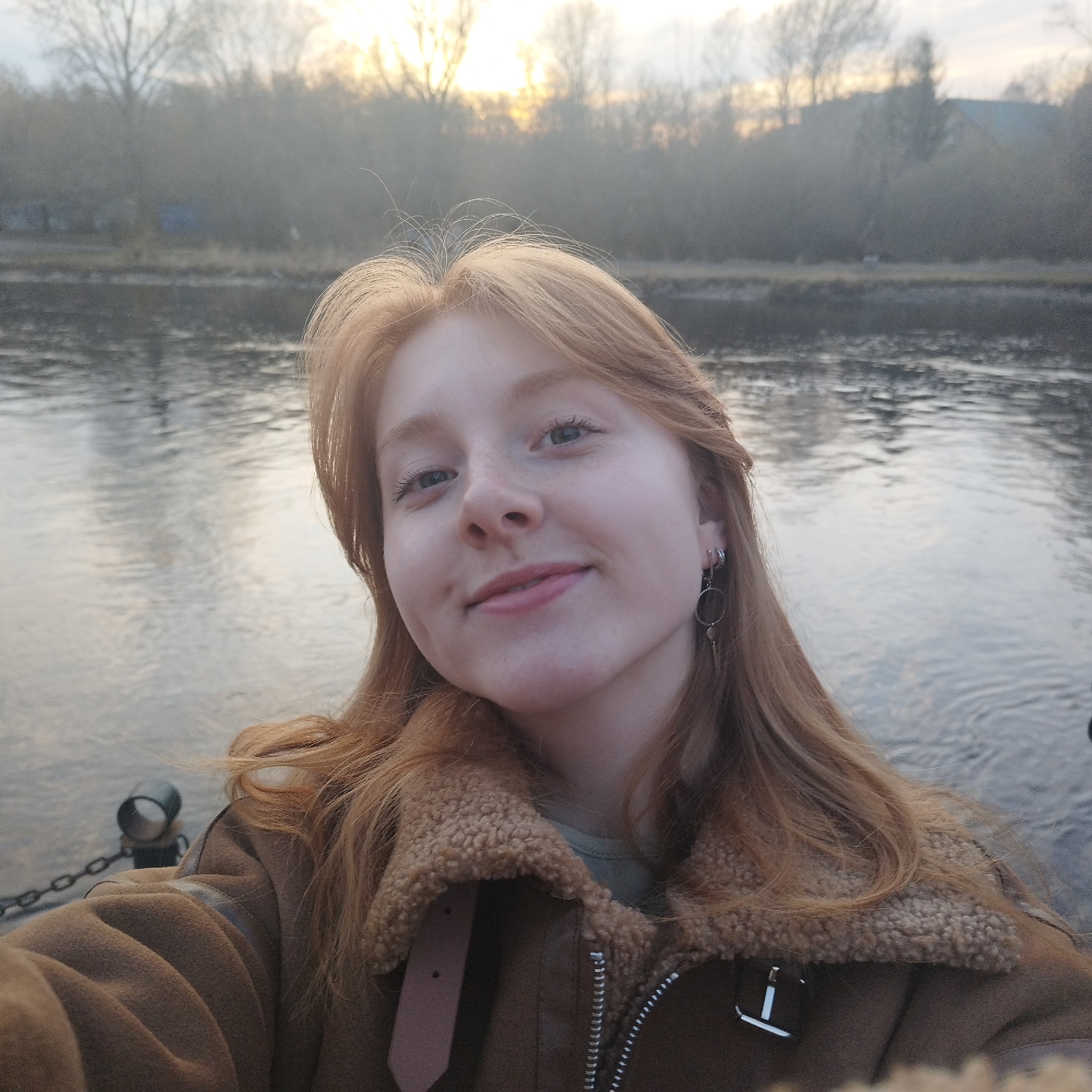 A selfie of a fair-skinned woman with reddish-blonde hair. The location appears to be a riverbank at sunset, potentially in Yekaterinburg, Russia. The woman wears a brown jacket with a shearling collar and dangling earrings. In the background is a river and trees are visible. A chain-link fence runs along the river's edge, with metal posts spaced out along it. The light suggests the time of day is either sunrise or sunset. The scene is somewhat foggy or misty.