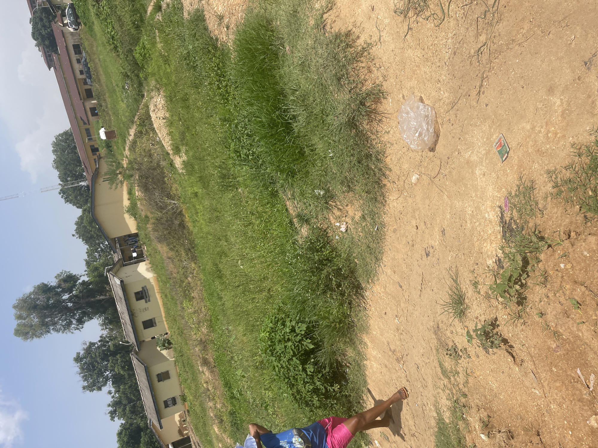 The image depicts an outdoor scene in what appears to be a somewhat neglected urban or semi-urban area, likely in Rafin Par, Nigeria, given the context. The foreground is dominated by a dirt path with scattered small green plants. To the right in the foreground, a crumpled plastic bag lies on the ground, and further to the right, a small, colorful packet, possibly candy or gum, is visible. In the middle ground, a person is partially visible on the left, wearing a blue t-shirt and pink shorts, walking on the dirt path. Behind the person, the ground slopes upwards, covered in thick, tall green grass. In the background, several low-lying, light-colored buildings with grey, sloped roofs are situated. Trees with dense foliage are visible behind the buildings and also scattered throughout the landscape. The sky is bright blue with some clouds. The overall impression is one of a sunny day in an area that shows signs of being a residential or institutional zone, with elements of both greenery and man-made structures.