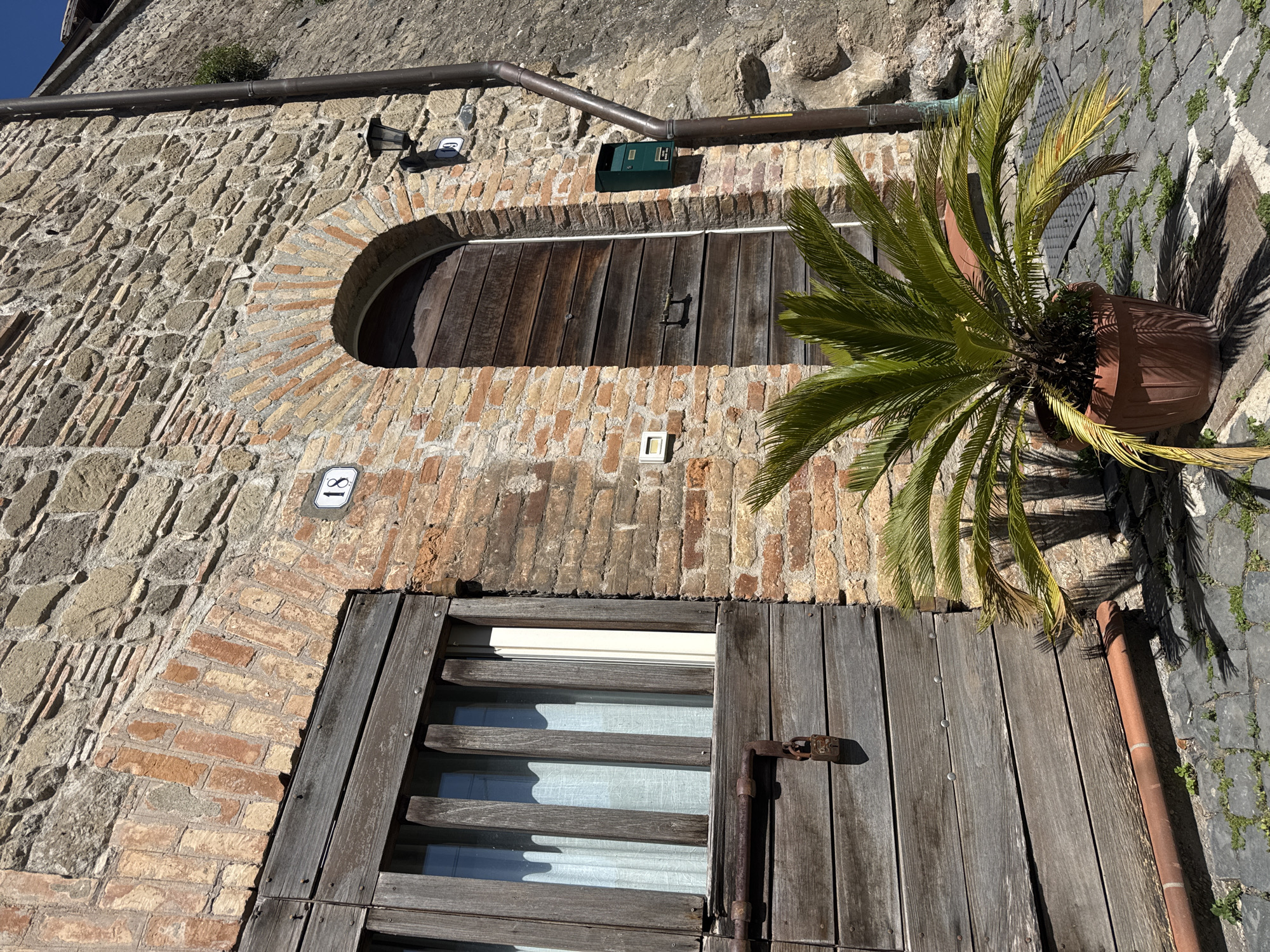 A low-angle, slightly tilted daytime shot shows the façade of a building in Castelnuovo di Porto, Italy. The building's exterior is constructed of rough-hewn stone and brick. The upper portion features a rounded wooden door framed by brick, above which is a green mailbox and a light fixture under a gutter. To the left of the door is a house number plaque displaying "17." Partially visible to the right of the door is a potted palm tree. Below the door, another set of wooden window shutters is closed and secured with a rusty latch. A house number plaque displaying "18" is above the shutters. The sidewalk is made of stone and features small patches of grass.