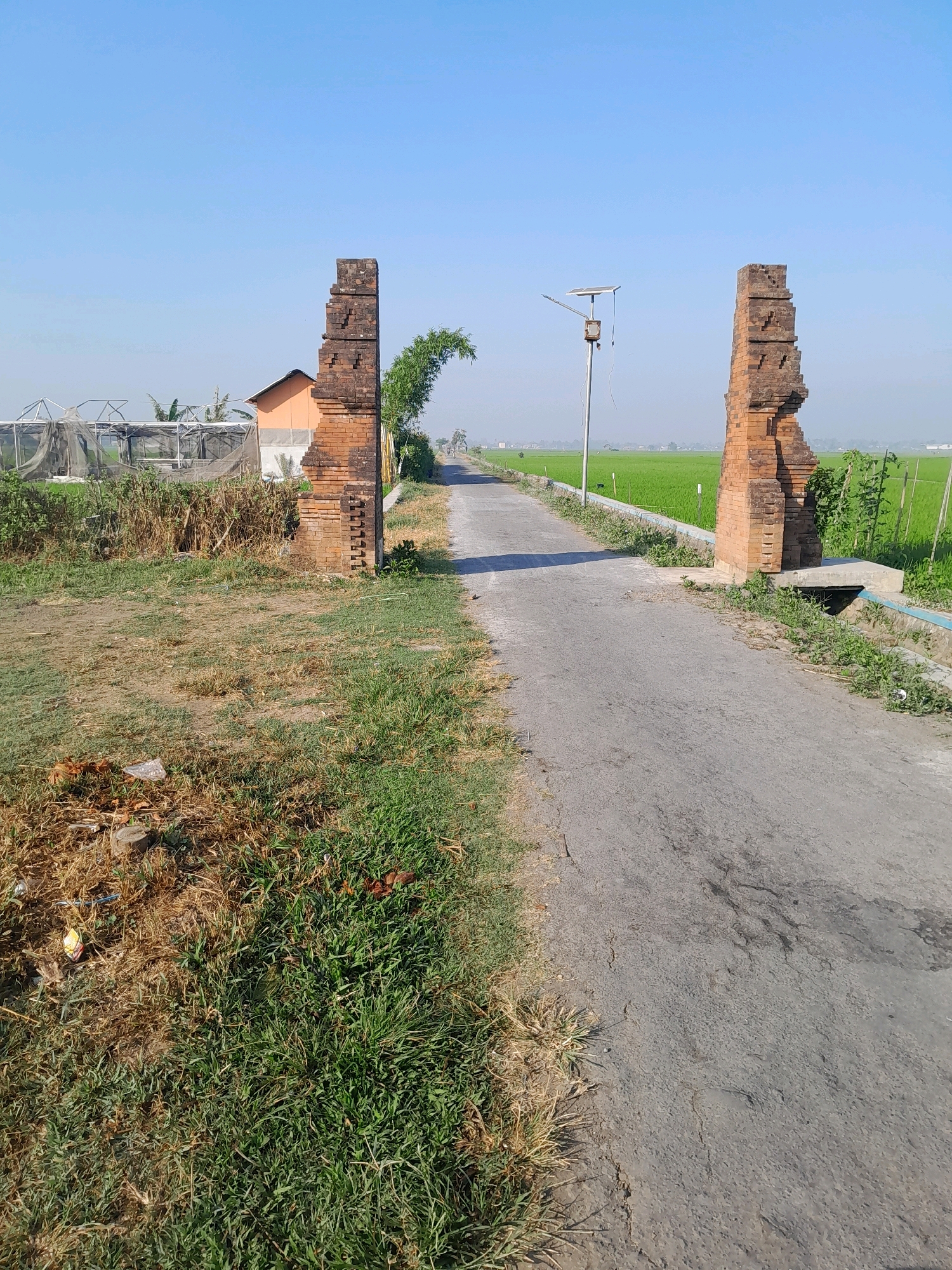 The image depicts an outdoor scene in Glanggang, Indonesia, featuring an aged brick gateway and an asphalt road stretching into the distance. The gateway, constructed from weathered red bricks, has a distinctly rustic and partially ruined appearance. To the left of the gateway, there is an orange-colored building with a light brown roof, surrounded by greenery and what appears to be a greenhouse structure. The right side of the gateway opens to a vast expanse of lush green rice paddies under a clear blue sky. A solar-powered street light is visible between the two brick pillars of the gateway. The road is bordered by grass and small plants, with some visible litter on the left side of the foreground. The overall environment suggests a rural or semi-rural setting in Glanggang, characterized by agricultural land and simple infrastructure.