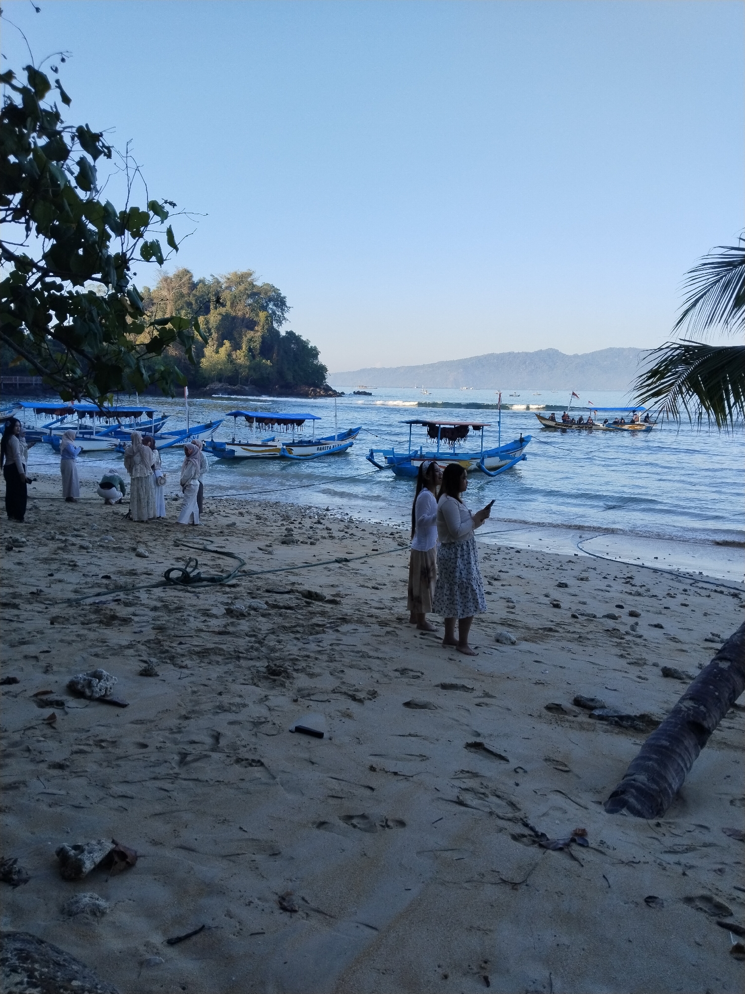 The image depicts a beach scene at Prigi, Indonesia, likely during the morning or late afternoon given the soft lighting. Several small, blue boats with canopy tops are anchored in the shallow, blue water near the shore. A group of people, some dressed in traditional modest attire, are visible on the sandy beach. Two women in the foreground are looking at a mobile phone. In the background, a tree-covered hill rises from the coastline, and distant mountains form the horizon under a clear, pale blue sky. The beach itself is sandy with scattered rocks and debris, and some footprints are visible in the sand. A fallen palm tree trunk is partially visible in the lower right corner of the frame.