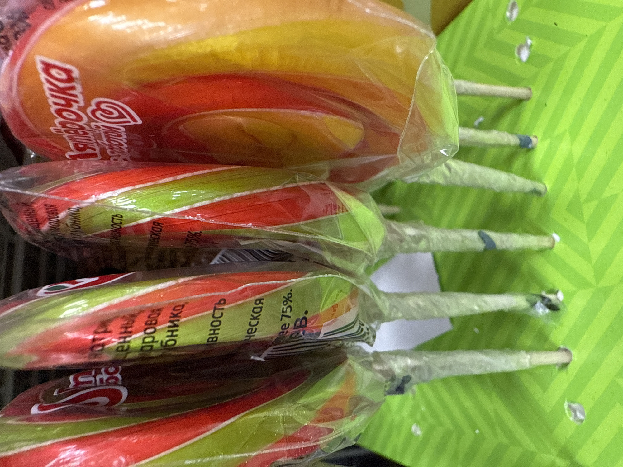 The image shows a close-up of several wrapped lollipops arranged in rows on a green surface. The lollipops are multicolored, with visible stripes of red, yellow, and green. The wrappers contain text in Russian. One of the lollipops has the text "Ялерочка" visible. The wrapper of another lollipop indicates "маровая клубника" and a detail that it contains "еская Mee 75%." The green surface is a perforated board where the lollipop sticks are inserted. The location is likely a retail setting, possibly in Votkinsk, Russia given the Cyrillic text on the wrappers.