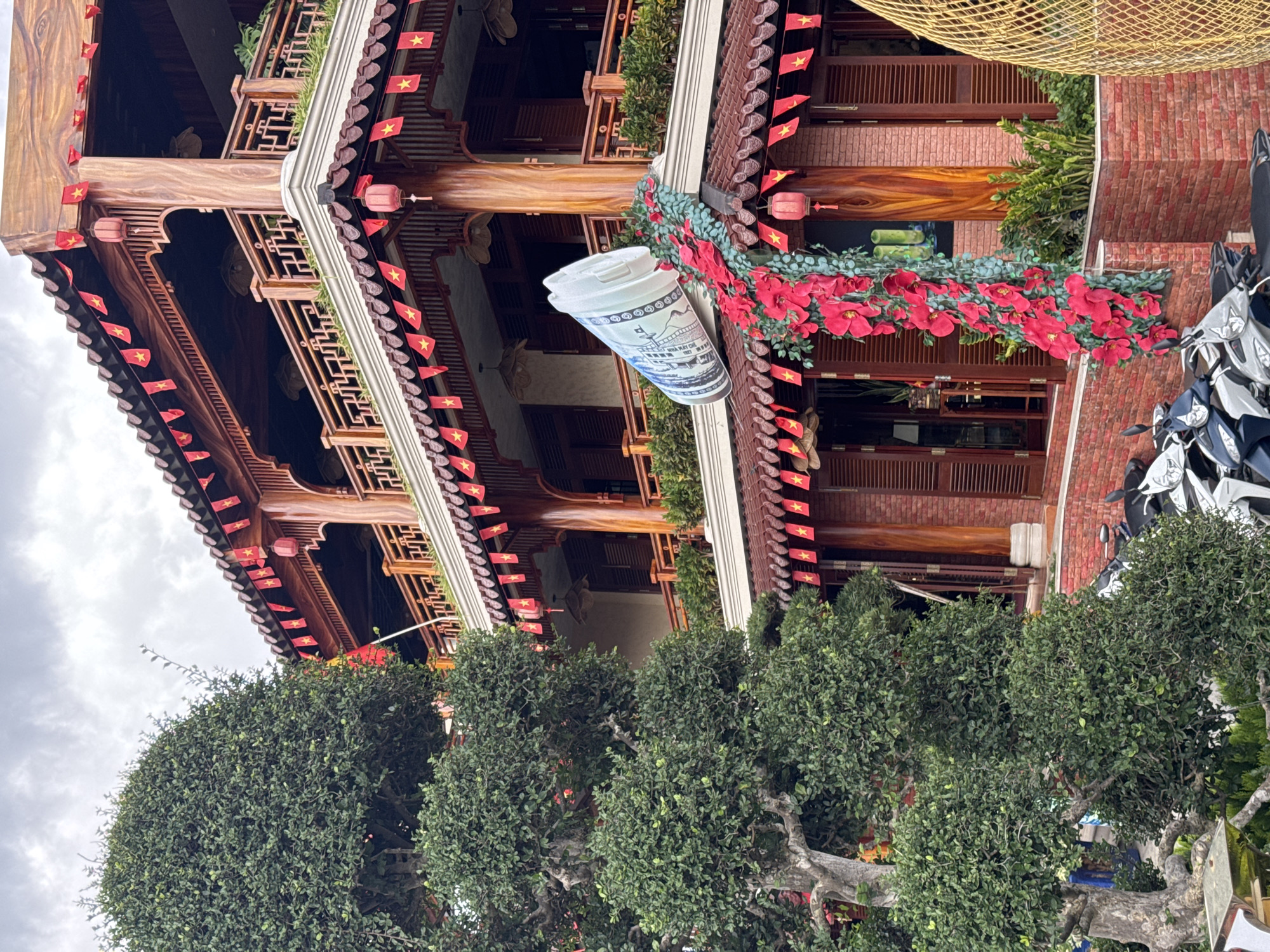The image is a low-angle, medium shot of a building in Da Lat, Vietnam. The building has a multi-tiered, East Asian architectural style, adorned with red Vietnamese flags and lanterns. A large, white styrofoam cup with blue detailing hangs prominently on the building's facade, surrounded by red flowers and greenery. Below the building are sculpted bushes. Parked to the side are several motorbikes. The sky is overcast.
