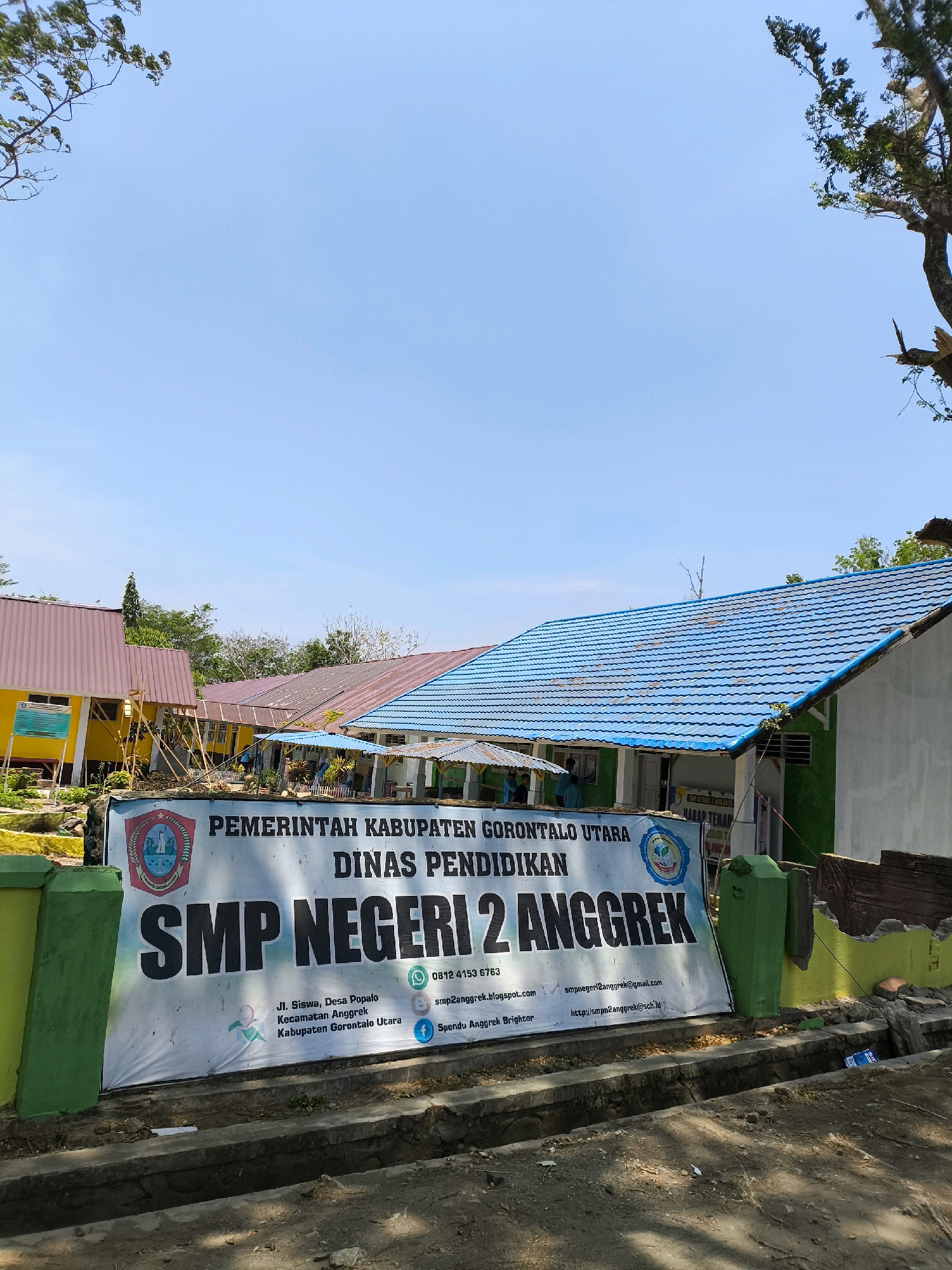 A wide, low-angle exterior shot captures a school building and its surroundings under a clear blue sky. In the foreground, a large banner is stretched across a green and yellow wall. The banner displays text in Indonesian, including the school's name, "SMP NEGERI 2 ANGGREK." Below this, an address is provided: "Jl. Siswa, Desa Popalo, Kecamatan Anggrek, Kabupaten Gorontalo Utara." Contact information such as a phone number (0812 4153 6783), an email address (smpn2anggrek@gmail.com), and a website (http://smpn2anggrek.sch.id) are also listed. The banner features various logos and icons, including a circular emblem with imagery of water and a palm tree, and social media icons for WhatsApp, Instagram, and Facebook.
Behind the banner, a portion of the school building is visible. It has a light green wall and a blue tiled roof. To the left of this building, another school structure with a pinkish-red roof is partially visible, painted yellow on its lower section. The school appears to be in a rural or semi-rural setting, with trees and vegetation surrounding the buildings. The ground in front of the banner is unpaved and shows signs of a ditch or drainage system. The overall scene suggests a daytime outdoor view of an educational institution.