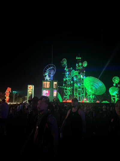 The image captures a nighttime scene in Indio, United States, likely at an outdoor event or festival. The foreground features several people amidst a large crowd, with individuals partially visible. The background is dominated by illuminated structures that resemble futuristic communication towers and satellite dishes, brightly lit in green and blue hues. There is a green beam of light shooting through the air above the structure to the right. Other structures are seen in the background with illuminated decorations. The overall setting suggests a vibrant and festive atmosphere.