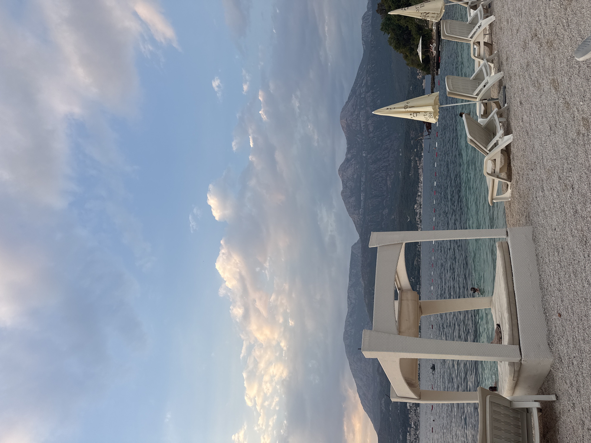 A wide shot of a beach at dusk. In the foreground, a white cabana bed with a cushioned surface is on the left. To the right of the cabana, there are several white lounge chairs, some with umbrellas. The beach is covered in small grey pebbles. The sea is a clear turquoise color, and in the distance, there are mountains. The sky is a pale blue with scattered white and pink clouds. There are a few people in the water near the cabana and further out in the sea. The overall scene depicts a serene beach setting during sunset.