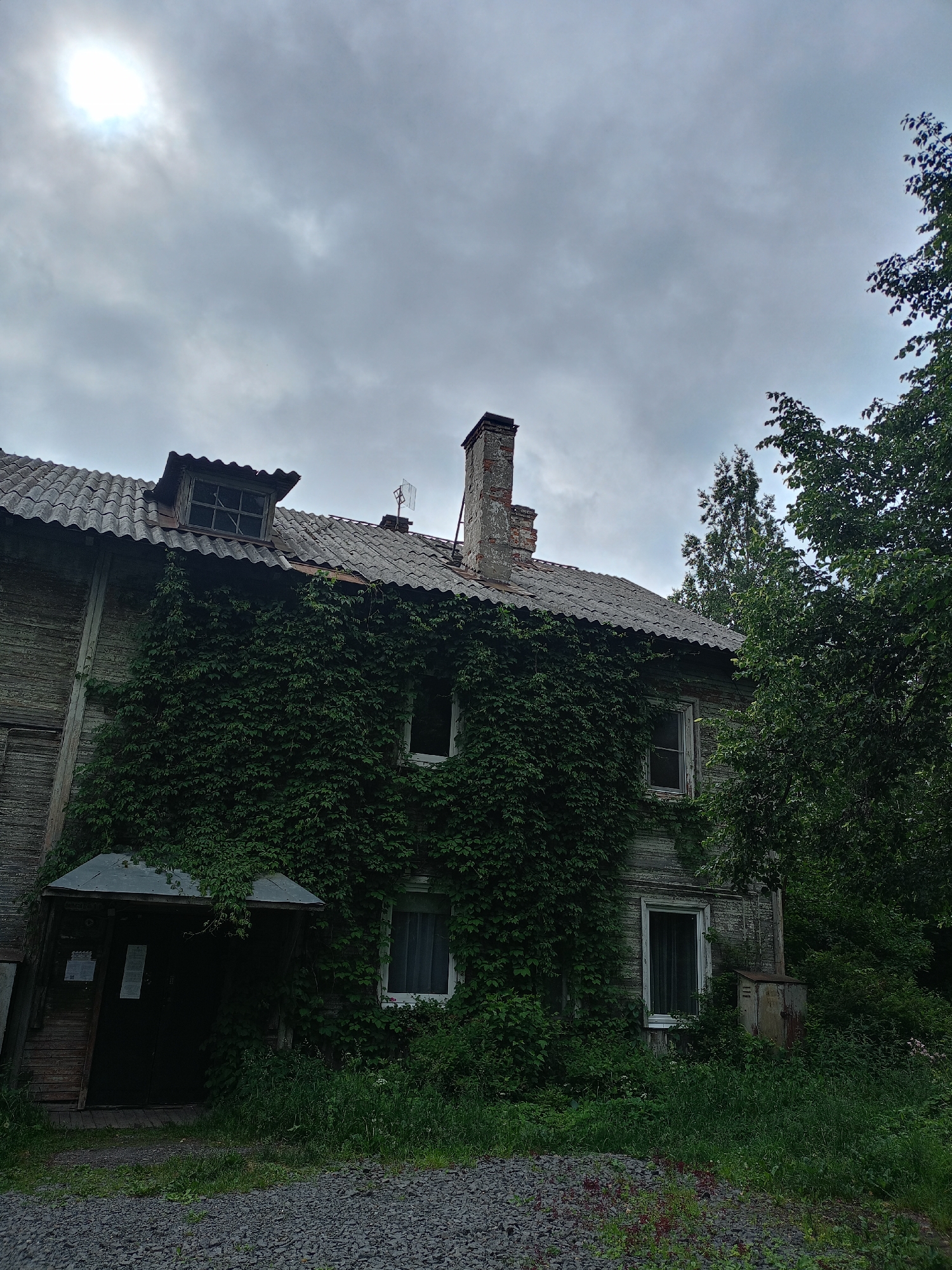 The image shows a dilapidated, two-story wooden building covered in ivy. The building has a corrugated roof, a brick chimney, and several windows. There is a small covered entrance with a dark door to the left of the building. The area in front of the building is covered in gravel and overgrown with grass and vegetation. Trees and foliage surround the building. The sky is overcast with a bright sun visible through the clouds. The scene appears to be in a rural or overgrown urban setting. There are no people visible in the image.
