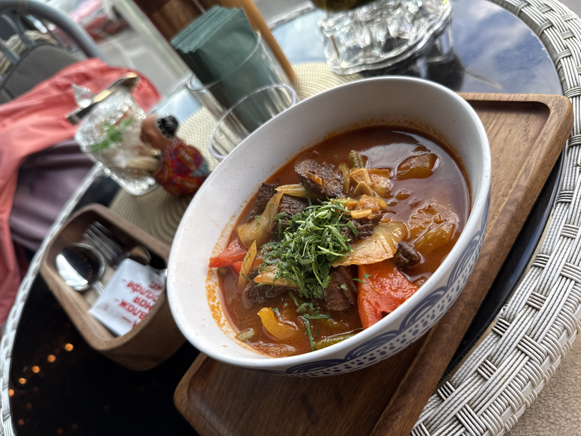 A close-up, high-angle shot shows a bowl of hearty stew filled with chunks of meat, red and yellow bell peppers, and onions, garnished with fresh herbs. The bowl, with its blue and white patterned rim, rests on a wooden tray. To the left, a small wooden container holds a spoon, a fork, and a white packet of sugar. In the background, out of focus, are glimpses of a table setting with a glass of water and a stack of green napkins. The image appears to be taken in a restaurant setting, possibly in Saint Petersburg, Russia, judging by the context provided. The lighting suggests it might be daytime, with reflections on the table surface indicating an indoor or covered outdoor environment.