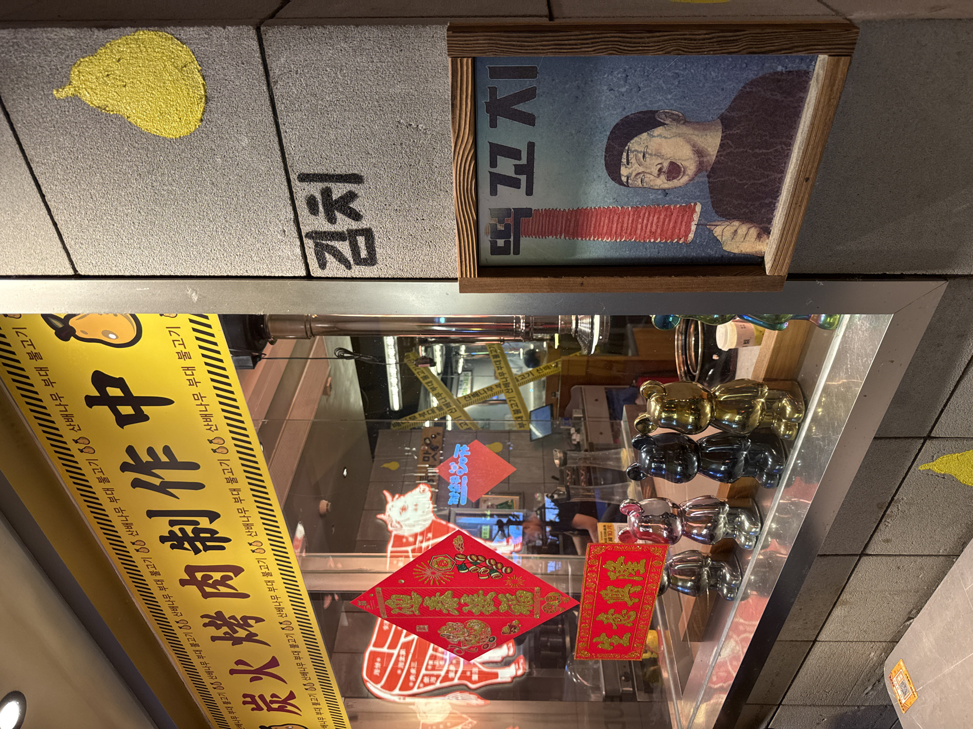 The image shows a shop front, possibly in the Haizhu District, China, indicated by Chinese characters and the overall visual style. There's a sign that reads "숯불돼지갈비 제작중" (Charcoal Pork Ribs in Production), followed by multiple occurrences of "산배나무 부대 불고기" (Sanbae Tree Stew Bulgogi). Above, "김치" (Kimchi) is written in Korean, and a framed picture depicts a person making "떡꼬치" (Tteokkochi). The shop window displays decorative items, including metallic bear figurines and red banners written in Chinese for the Lunar New Year, wishing for prosperity. There's also a diagram of meat cuts on display. The shop seems to specialize in Korean-style BBQ, particularly bulgogi, and the Lunar New Year decorations suggest a festive atmosphere. Caution tape is affixed above the shop window. A yellow spot is painted on the wall.