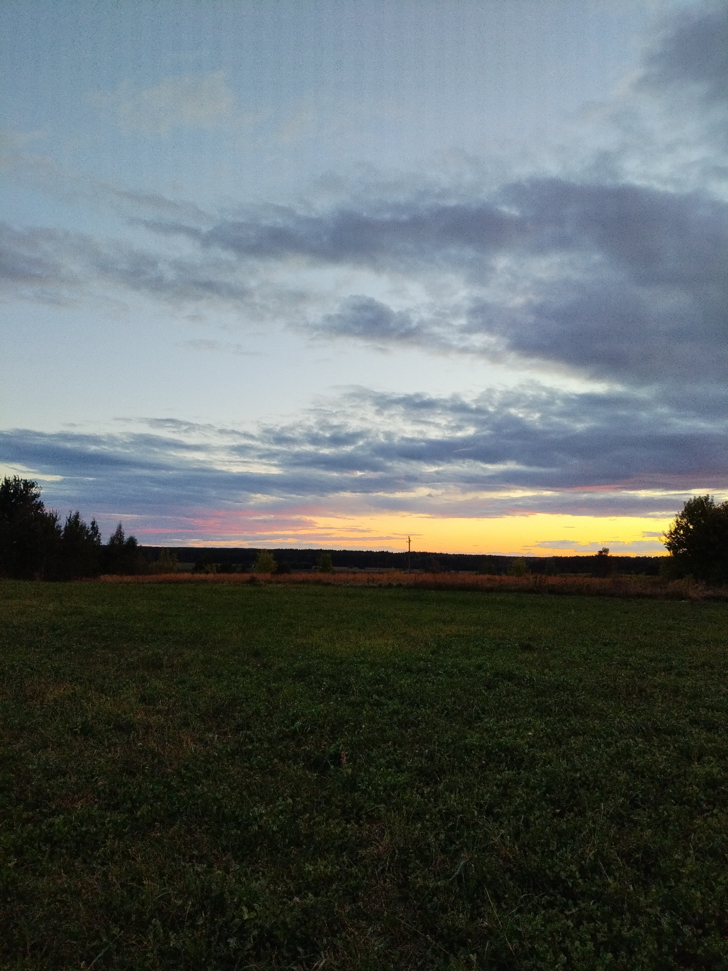 This is a landscape photograph taken at dusk in the Lan'kaŭski sel'ski Savet, Belarus. The image features a wide, grassy field in the foreground, with a dark line of trees forming the horizon. Above the trees, a dramatic sky displays layers of clouds, with a vibrant band of yellow and pink light from the setting sun visible just above the treeline. The overall impression is one of rural tranquility and natural beauty at the end of the day.