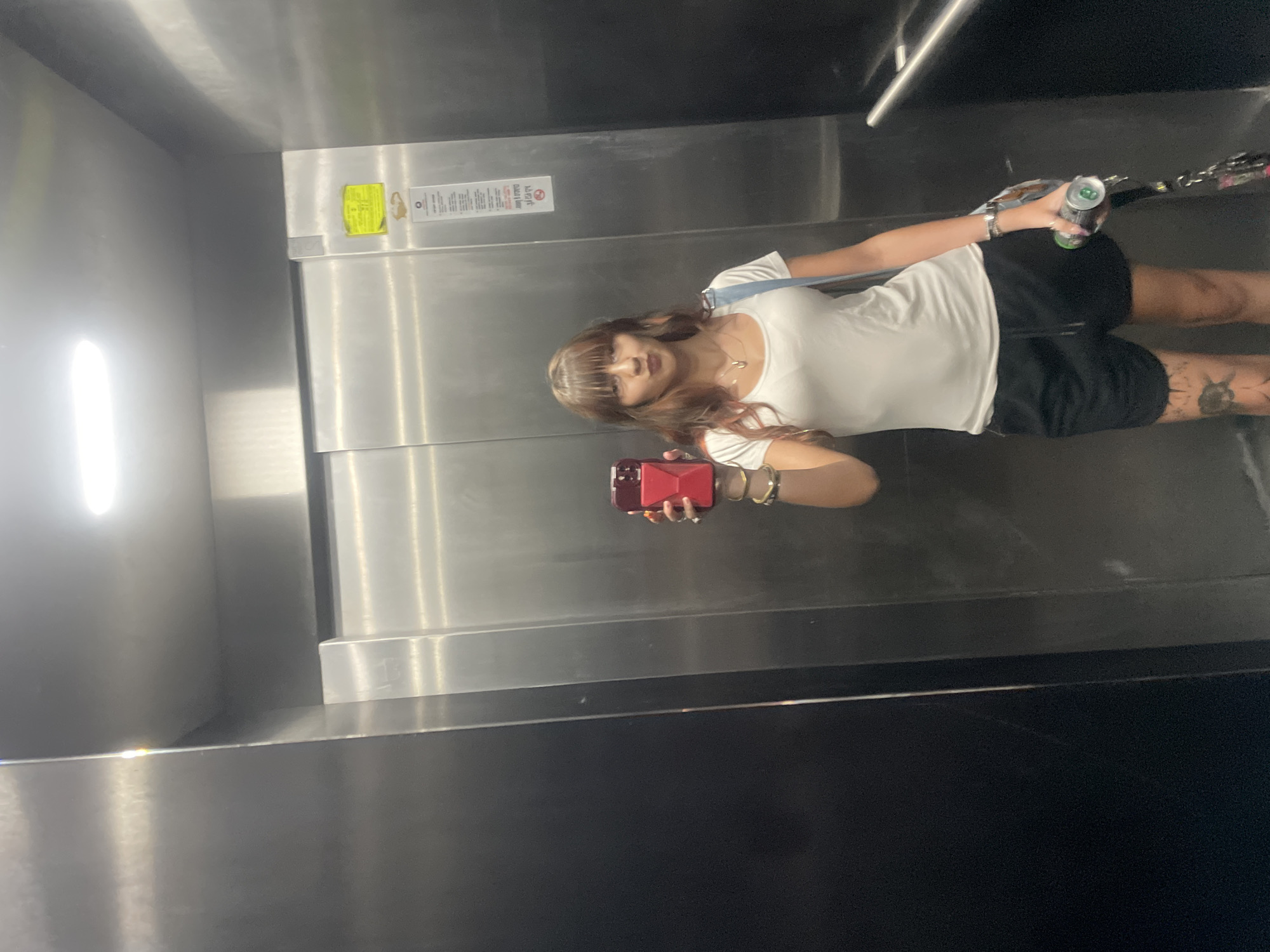 A young woman with brown hair and bangs stands in an elevator, holding a red cellphone in her left hand. She is wearing a white t-shirt, black shorts, and a light blue strap across her chest. Her right hand holds a green can. She is looking towards her cellphone, taking a mirror selfie. The elevator has stainless steel walls and a light fixture on the ceiling. A yellow sticker is visible on the right wall.
