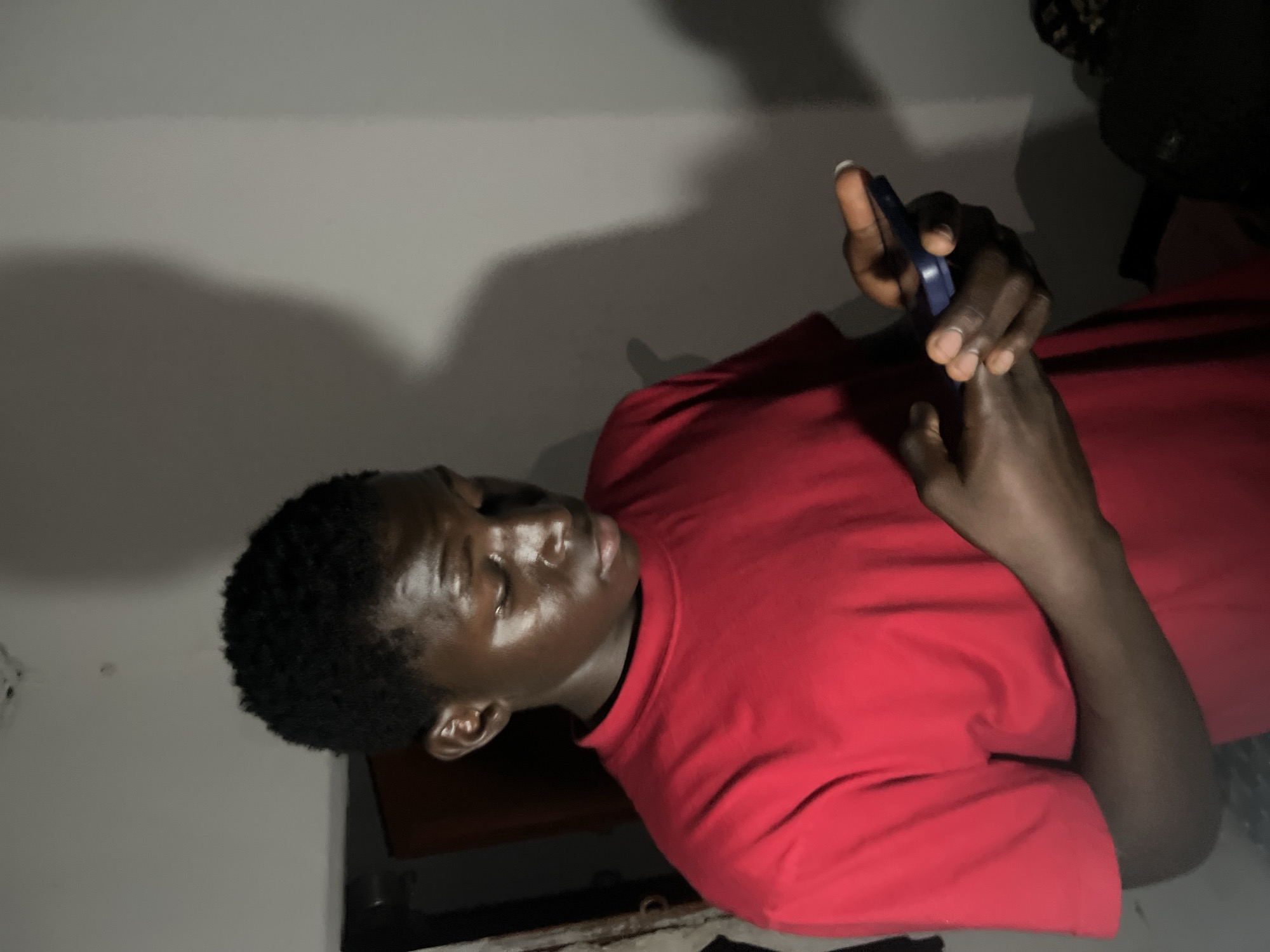 The image shows a young Black male wearing a red t-shirt, holding a dark blue smartphone in his hands and looking down at it. He is standing in what appears to be an indoor setting. The lighting is dim, casting a shadow of the man on the wall behind him. The background is plain and light-colored, with some indistinct objects visible to the left and right. There are no visible indications of the specific location within Agwala, Nigeria. The image captures a moment of personal engagement with technology, devoid of any outward interaction with others or the surrounding environment.