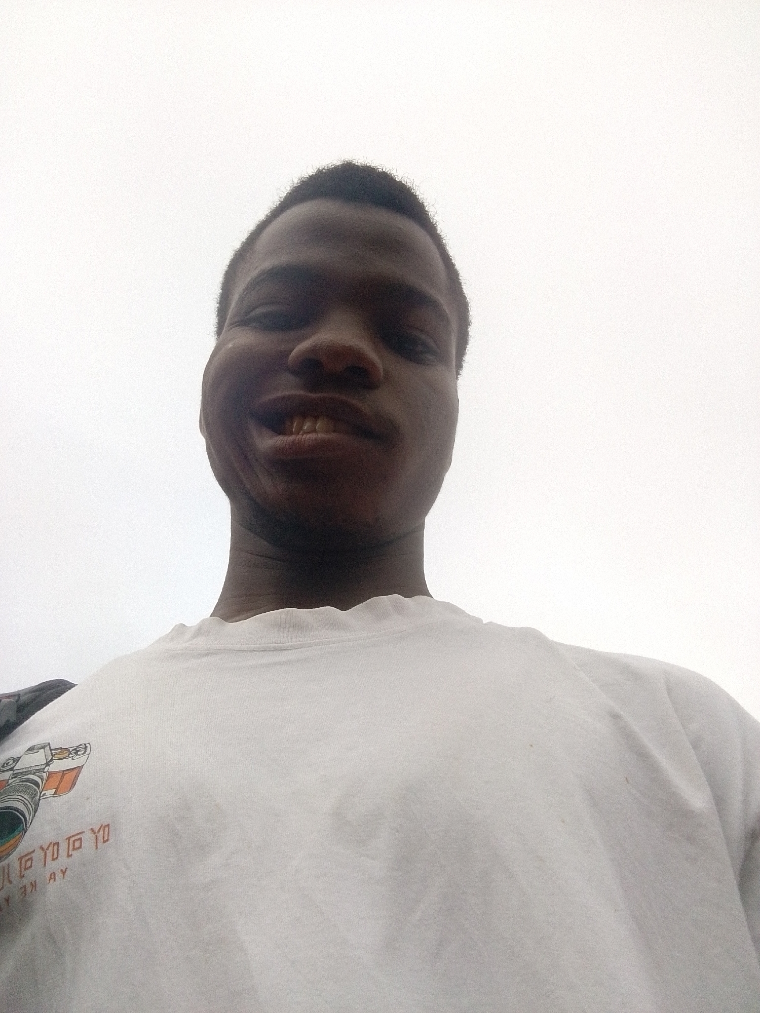 A selfie of a Black male with short, dark hair wearing a white t-shirt is centered against a bright, overcast sky. A camera graphic is visible on the t-shirt. The location is Unknown, Unknown.