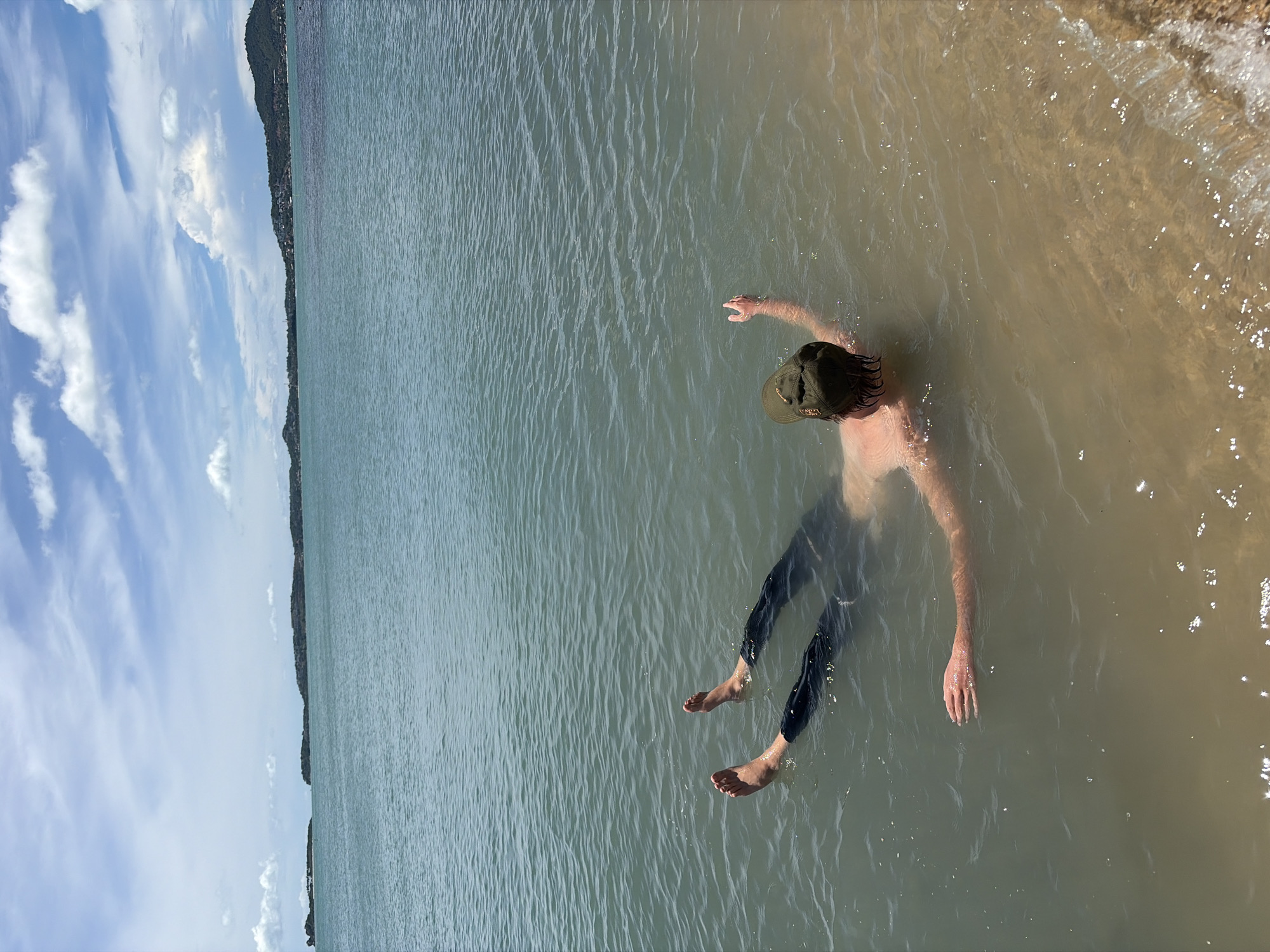 A person is floating on their back in shallow, clear ocean water near the shore in Baan Bo Phut, Thailand. The person is wearing a green baseball cap and dark pants. Their arms are extended upwards and outwards, and their legs are slightly bent. The water is calm with small ripples. In the background, there is a coastline with green hills and a blue sky with scattered white clouds. The sandy bottom is visible beneath the water.