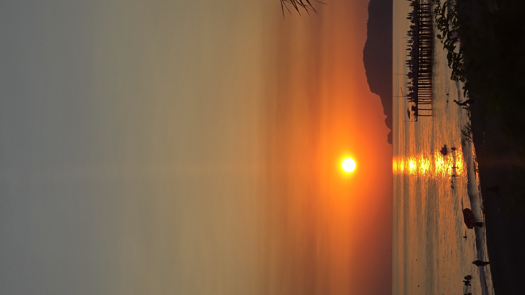 A vertical, low-angle photograph captures a vibrant sunset over the sea in Alanya, Turkey. The sun, a bright orange orb, is positioned in the lower half of the frame, casting a shimmering golden reflection on the calm water. A silhouetted pier with numerous umbrellas and people extends into the sea from the right side of the image. In the distance, the dark silhouette of a mountain range lines the horizon. The beach in the foreground is dark and indistinct, with several people visible wading in the shallow water or strolling along the shore. The sky transitions from a pale grey at the top to a gradient of orange and yellow as it nears the horizon.