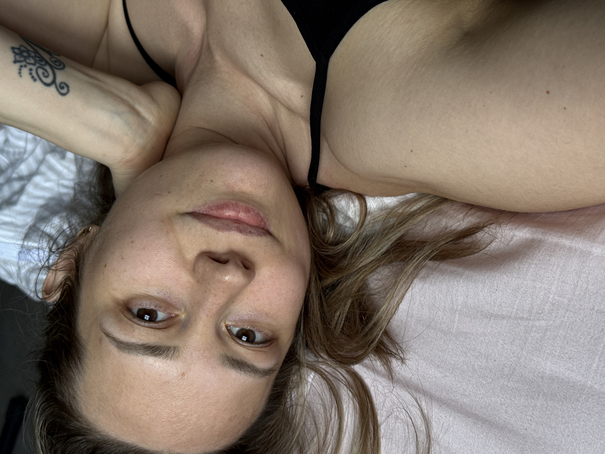 A medium shot depicts a woman lying on a light-colored surface, possibly a bed, from an overhead perspective. Her head is oriented towards the bottom of the frame. She has fair skin, brown eyes, and light brown hair that is loosely scattered around her head. A tattoo is visible on her left arm. She is wearing a black garment with thin straps. The location is not possible to determine from the image.