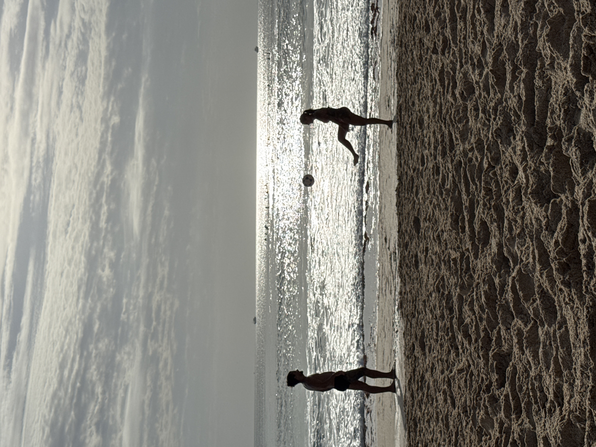 Two individuals, silhouetted against a bright, hazy sky and shimmering ocean, are on a sandy beach in Kuta Selatan, Indonesia. On the left, a male with his hair tied back stands facing the ocean. On the right, a female is in the process of kicking a soccer ball, which is airborne in front of her. The beach is composed of light brown sand with visible textures. The ocean stretches to the horizon, with a bright reflection of the sun on its surface. A few distant boats are visible on the water. The sky is filled with layered, wispy clouds.