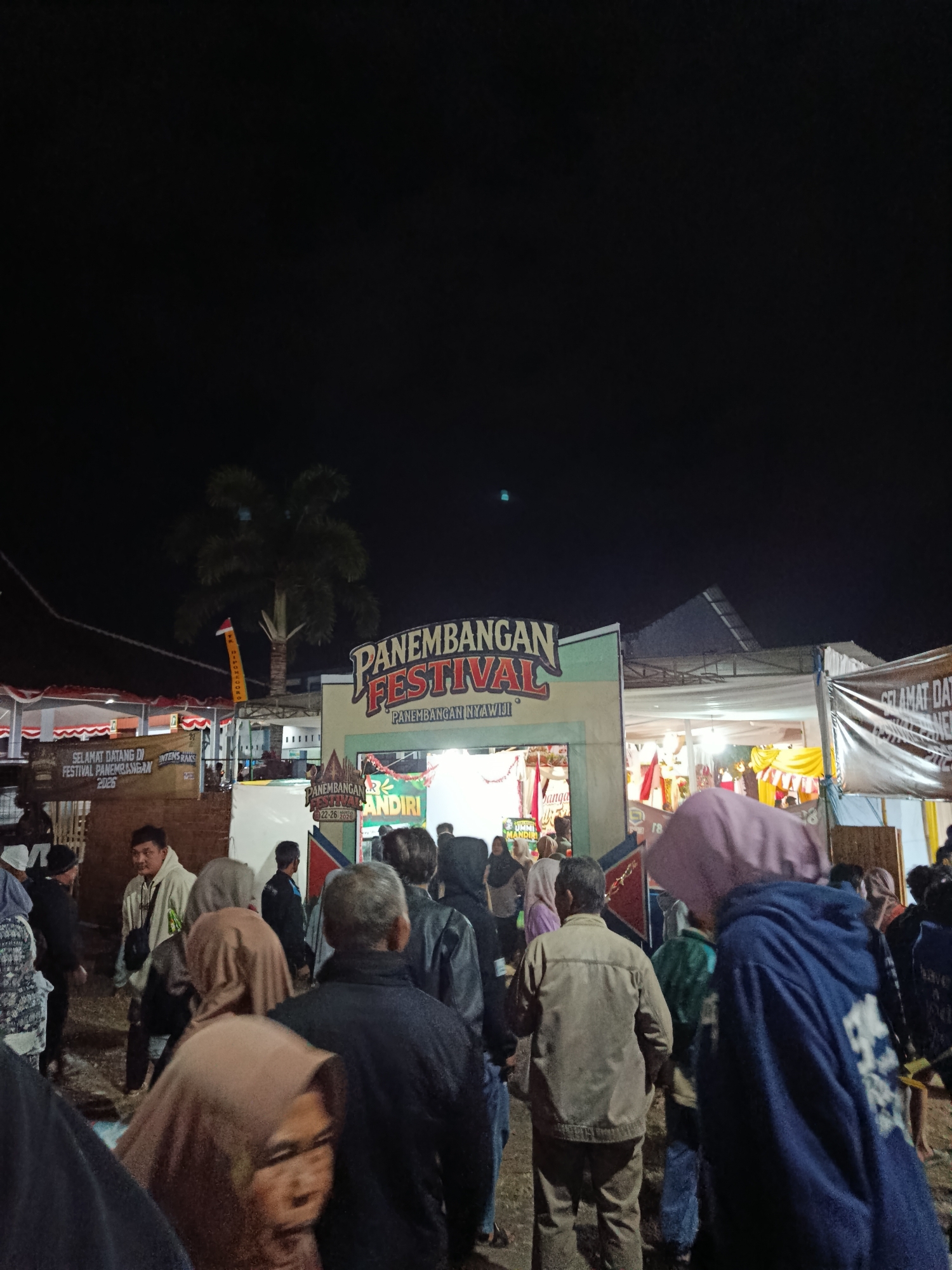 The image captures an outdoor nighttime scene at the Panembangan Festival in Panembangan, Indonesia. A crowd of people is gathered, seemingly queuing to enter an arched gateway decorated with colorful banners and text. The main banner above the arch reads "PANEMBANGAN FESTIVAL" in large, stylized letters, with smaller text beneath it. Other banners are visible to the left and right of the arch, with one on the left welcoming visitors to the festival. A tall palm tree is visible in the background, silhouetted against the night sky, along with some buildings and stalls that are illuminated by artificial lighting. The ground appears to be a dirt or gravel surface. People are dressed in casual attire, with some wearing headscarves. The overall atmosphere suggests a lively event or celebration.