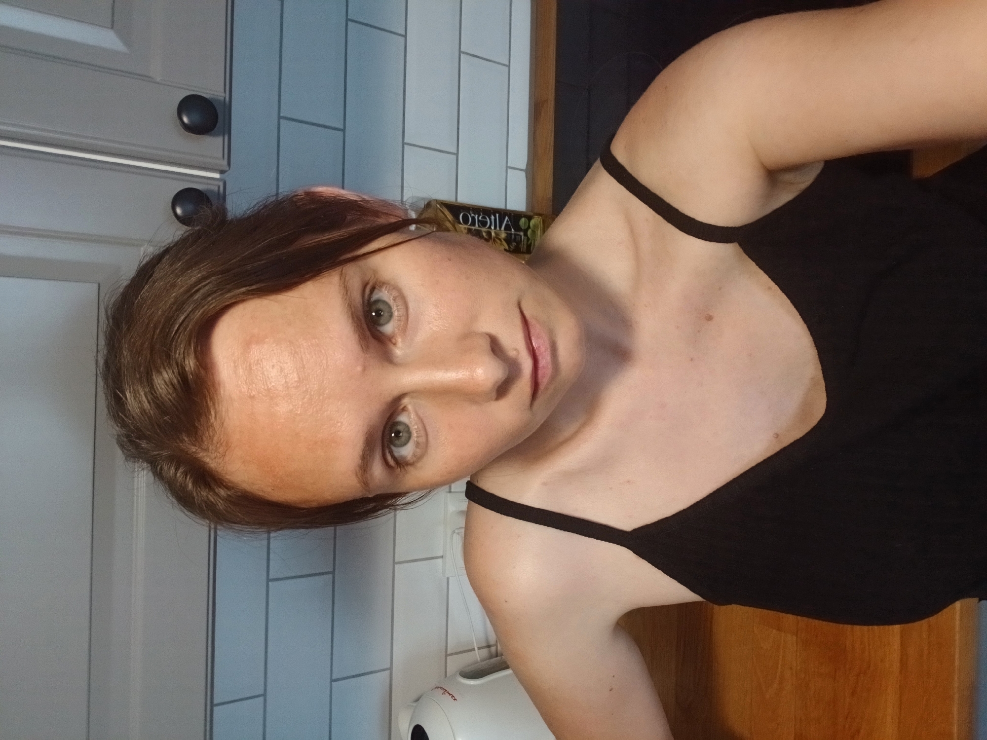 A woman with brown hair and green eyes is taking a selfie in a kitchen. She is wearing a black tank top. Behind her, there are white subway tiles on the wall and gray cabinets with black knobs. A white electric kettle is visible on the left side of the image. There is also a bottle of olive oil on a wooden countertop on the right side of the image.