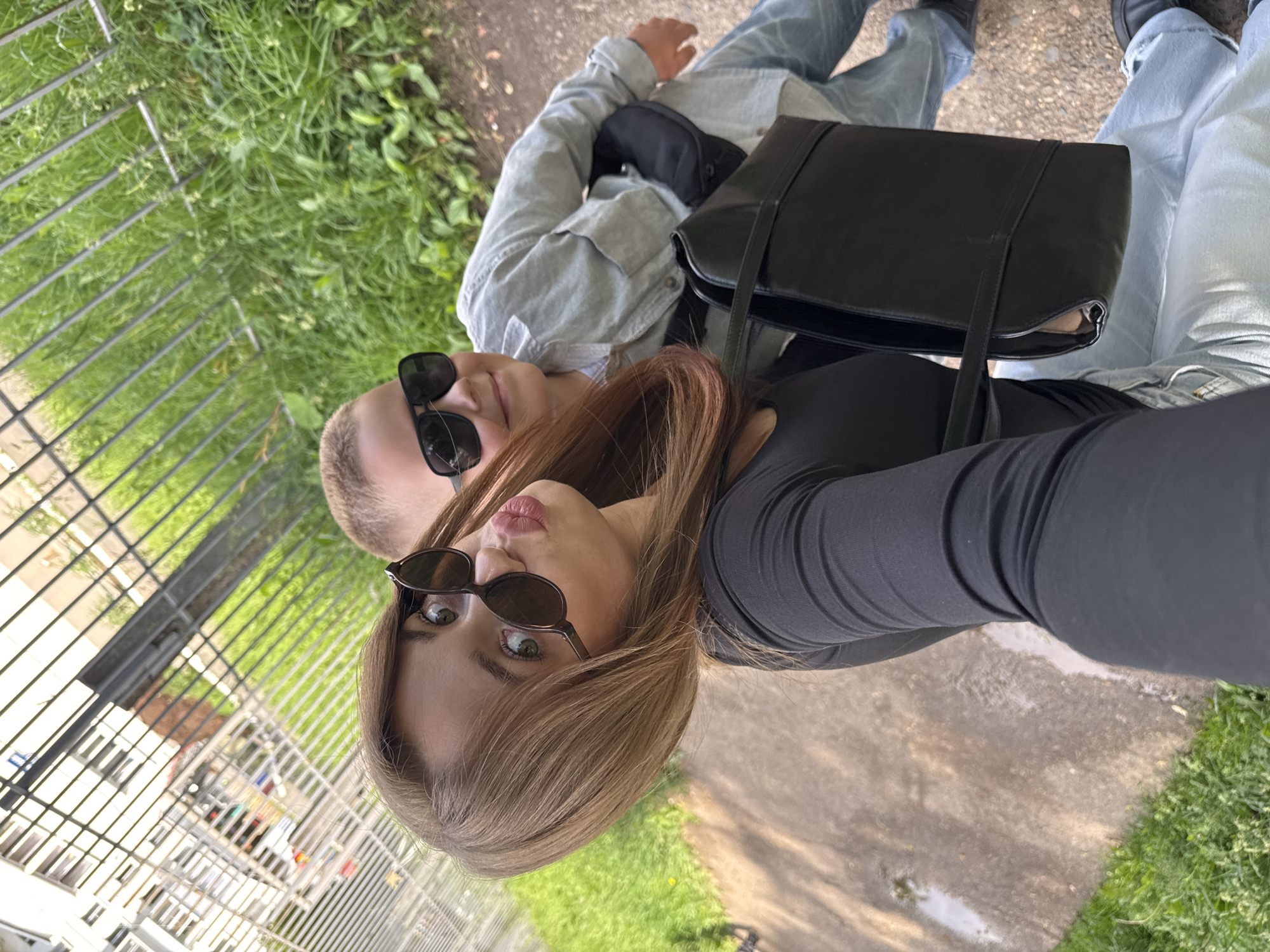 In a high-angle, selfie-style shot, a young woman and a young boy are outdoors, with a metal fence and green grass in the background, suggesting an urban park or residential area, possibly in Chaikovsky, Russia. The woman, positioned on the left, wears a black long-sleeved top and sunglasses, her lips pursed as if for a kiss. She has light brown hair with subtle pink highlights. Beside her, to the right, the boy is also wearing sunglasses and a light gray button-up shirt. He has short blond hair and a slight smile. The woman is holding a black handbag with a structured rectangular shape and a single strap. The boy appears to be seated behind the woman. The ground is a mix of concrete and dirt, with patches of grass. The overall lighting suggests a daytime scene.