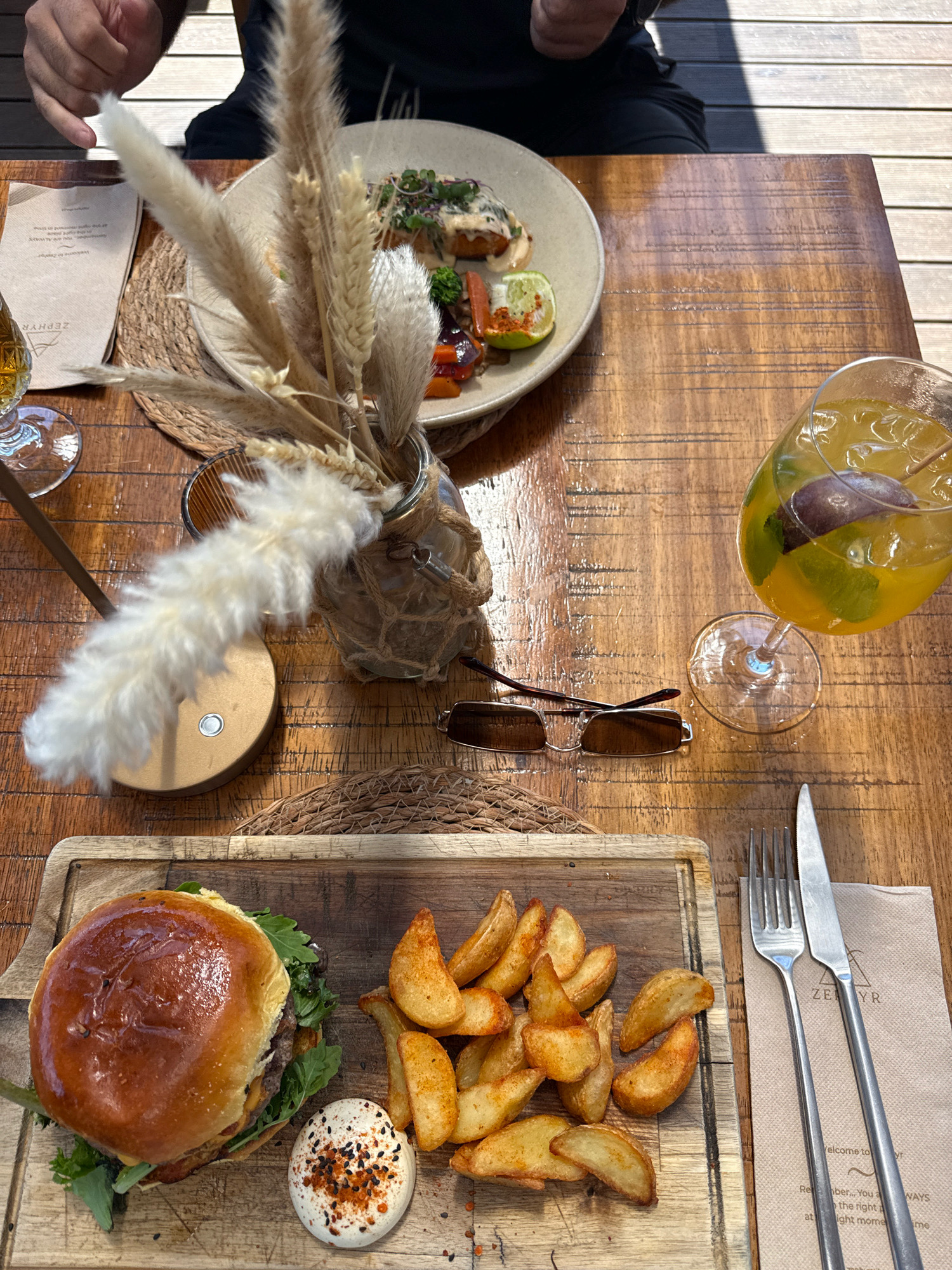 The image shows a wooden table set with a meal. In the foreground, a burger and potato wedges are served on a wooden board. Next to them are a fork and knife on a paper napkin with the logo "ZEPHYR". A drink in a wine glass is visible to the right, containing a yellow liquid and a dark berry. In the background, a plate of food with greens and sauce is partially visible, as well as a vase of dried flowers. A pair of sunglasses rests on the table. The scene appears to be outdoors, with sunlight casting shadows on the table, suggesting a restaurant or cafe setting. The presence of the "ZEPHYR" logo on the napkin indicates the meal is from the restaurant Zephyr, located in Vila Nova de Gaia, Portugal.