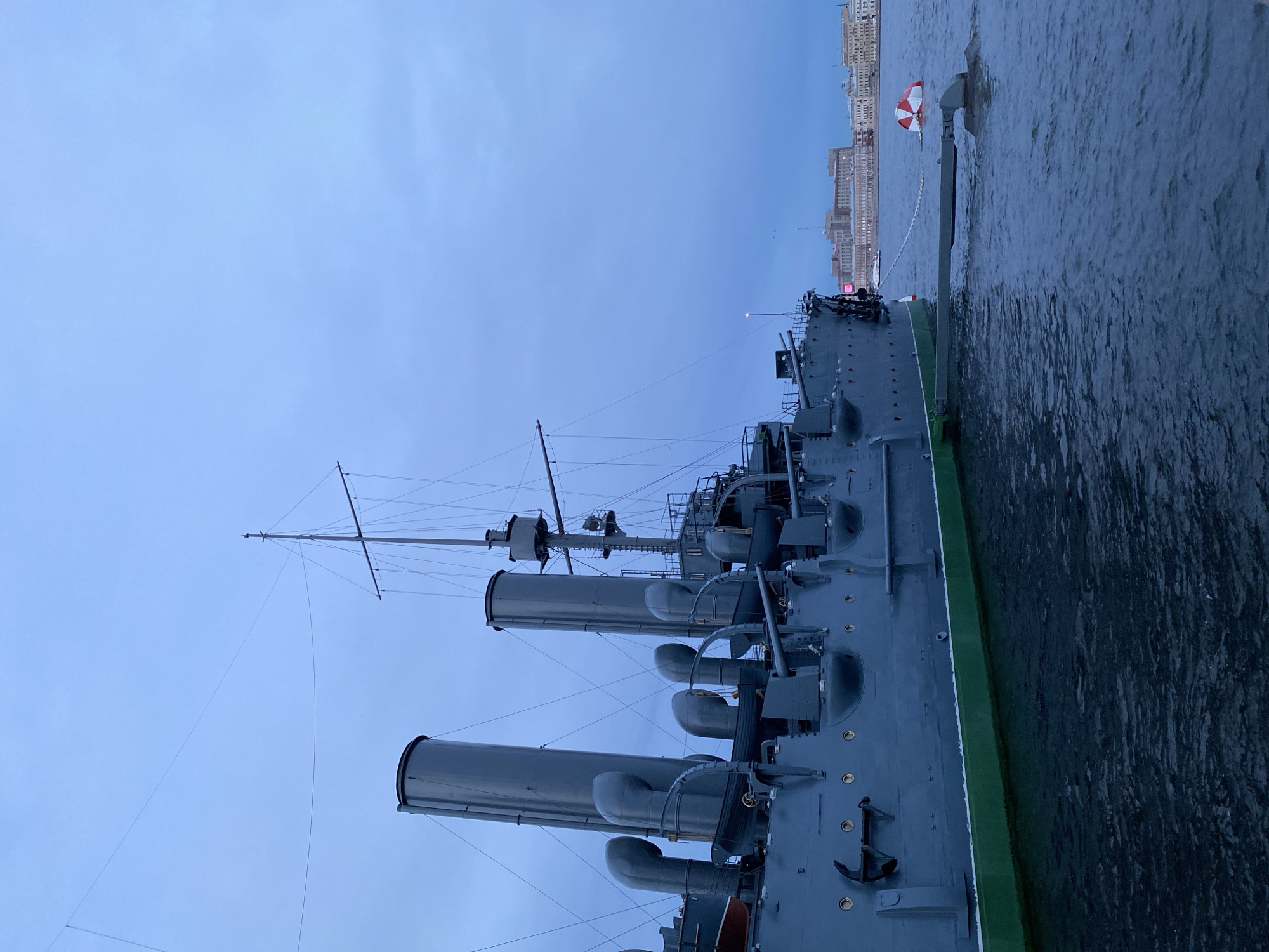 The image shows the cruiser Aurora, a museum ship moored on the Neva River in Saint Petersburg, Russia. The ship, painted a dark gray with a green waterline, is positioned on the left side of the frame. Its prominent features include multiple smokestacks, large pipes connecting to them, cannons mounted on the deck, and lifeboats. In the foreground, the dark, rippling water of the Neva River is visible. To the right of the ship, a red and white buoy is floating in the water. In the background, across the river, is a cityscape with multiple buildings, including architectural structures that suggest historical significance, consistent with the architecture found in Saint Petersburg. The sky is overcast and a pale blue, indicating either early morning or late evening. There are no people visible in the image.