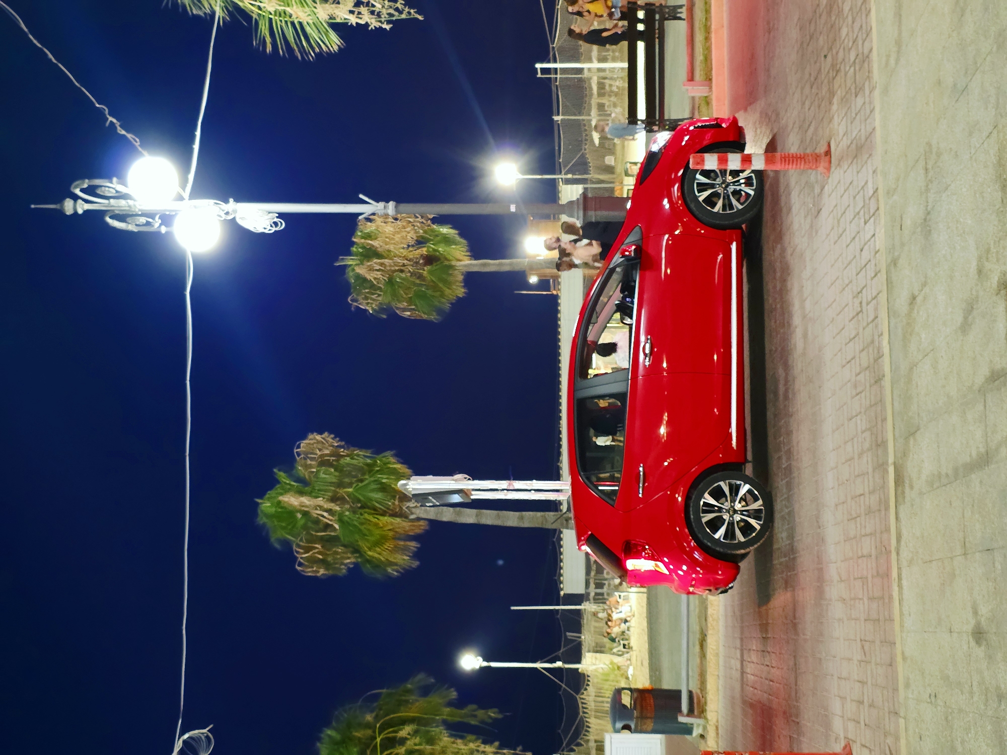 A red Kia Picanto is parked at night on a paved street lined with palm trees. In the background, a lamppost illuminates a group of people standing near a beachside establishment. The scene appears to be along the Finikoudes promenade in Larnaca, Cyprus, known for its palm-lined beachfront and lively atmosphere. The car's headlights are on, casting a glow on the street. People are visible in and around the car, suggesting activity or a stop. The overall impression is of a warm evening in a coastal city.