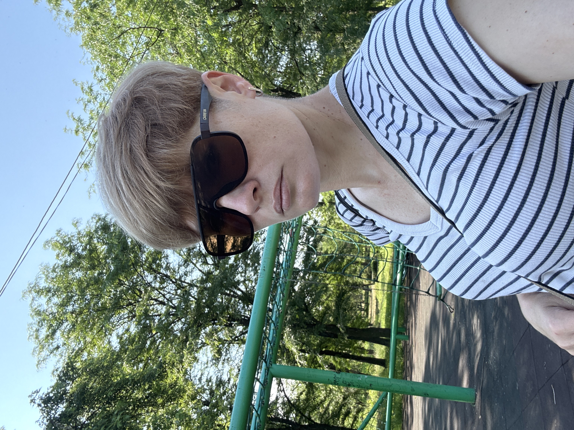 A woman with short, blonde hair and sunglasses takes a selfie outdoors. She is wearing a black and white striped t-shirt. She is standing in front of a green metal playground structure. The background consists of trees and a bright blue sky.