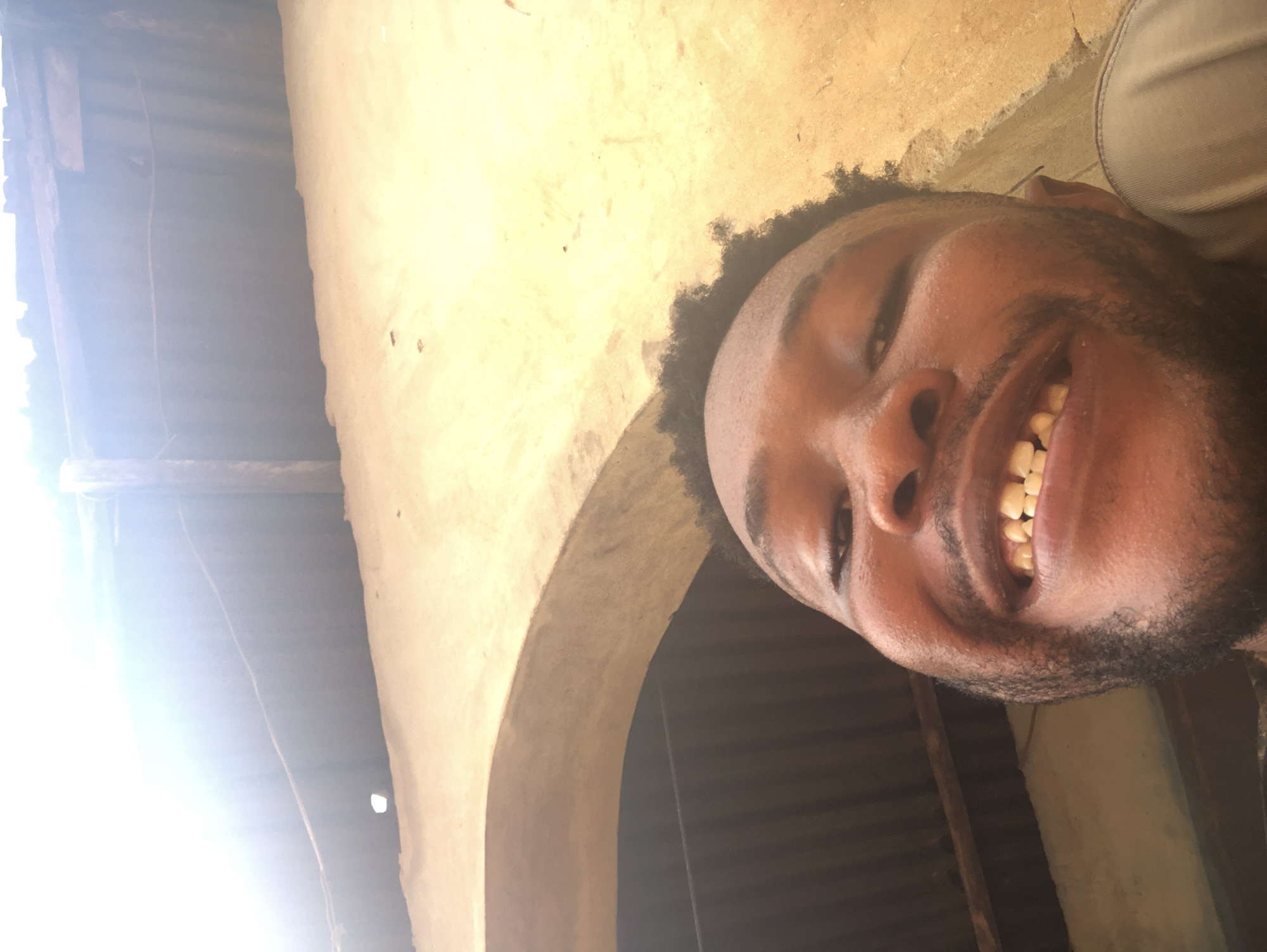 A low-angle shot shows a man smiling, likely taken from his own perspective. He has dark skin, a close-cropped beard and hair, and is looking downward into the camera. His teeth are visible in his broad smile.
The background is somewhat blurred and suggests an indoor or covered area. There's a textured, light-colored wall partially visible, possibly made of mud or stucco. Behind the wall, there are dark, corrugated metal sheets, perhaps forming a roof or enclosure. An archway is visible to the side of the man's face.
The location is described as the city of Unknown, Unknown.