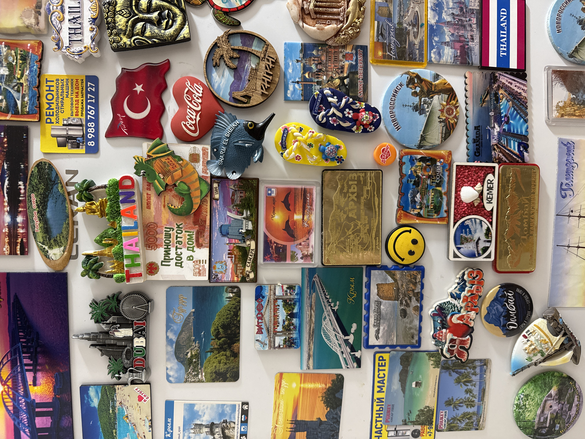 The image displays a close-up, high-angle view of a refrigerator door adorned with a collection of novelty magnets. The magnets feature various landmarks and designs, primarily representing tourist destinations. Prominently featured are magnets depicting Dubai, Thailand, Crimea, and St. Petersburg. Other magnets showcase a Turkish flag, a Coca-Cola logo, a cartoon dragon, a smiling face, and images of beaches, bridges, and historical buildings. The overall impression is one of a decorative display of souvenirs, suggesting a personal collection. There is no discernible information within the image that can identify the specific location as Mikhaylovsk, Russia.