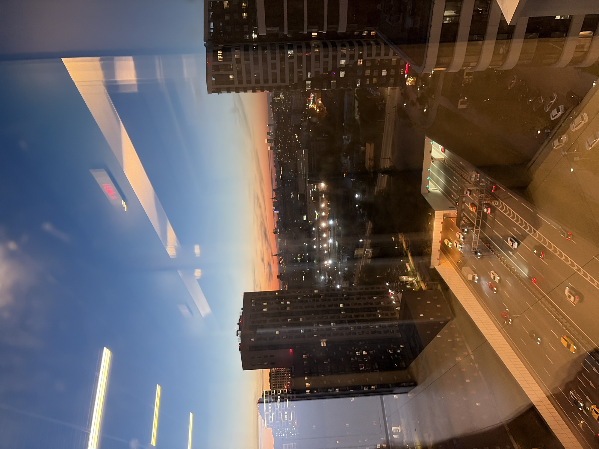A high-angle shot captures a city scene at dusk, viewed through a glass surface reflecting lights and the sky. The foreground shows a multi-lane highway with active traffic, including cars and trucks, leading into a tunnel entrance illuminated by green lights. Adjacent to the highway, a tall, modern building with numerous illuminated windows dominates the mid-ground, casting reflections on the glass. In the distance, the cityscape stretches out, dotted with lights and fading into the twilight sky, which displays a gradient of blue and orange hues. The overall impression is an urban environment at the transition from day to night, with artificial lights illuminating the infrastructure and buildings.