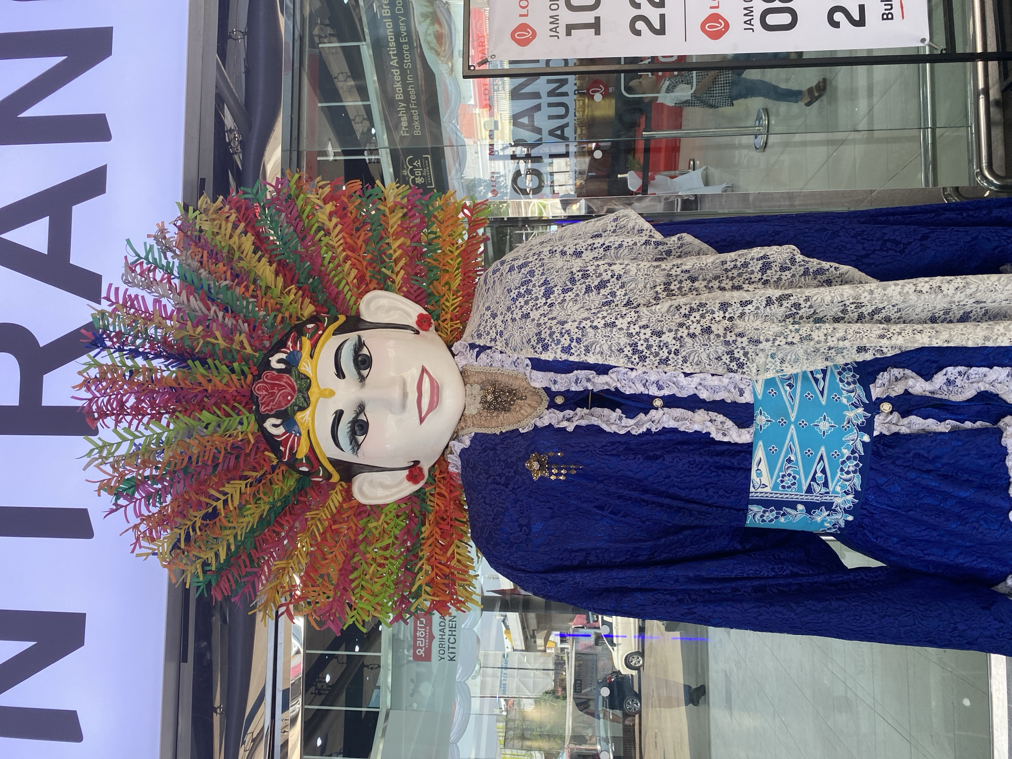 A full-body, eye-level, outdoor shot depicts a large, traditional Indonesian Wayang Golek puppet standing in front of a glass-fronted building. The puppet is dressed in a vibrant royal blue lace garment, adorned with a white lace shawl draped over its shoulder. A decorative blue and white sash is cinched around its waist. The puppet's face is white with painted-on features, including bright blue eyes and red lips. It wears an elaborate headdress made of colorful, feathery paper in shades of red, orange, yellow, green, and blue. The building behind the puppet displays a "GRAND LAUNCH" sign, indicating its presence in North Jakarta, Indonesia. Through the glass, reflections of vehicles and people are visible, suggesting a busy urban environment. The lighting is bright, indicating it is daytime.
