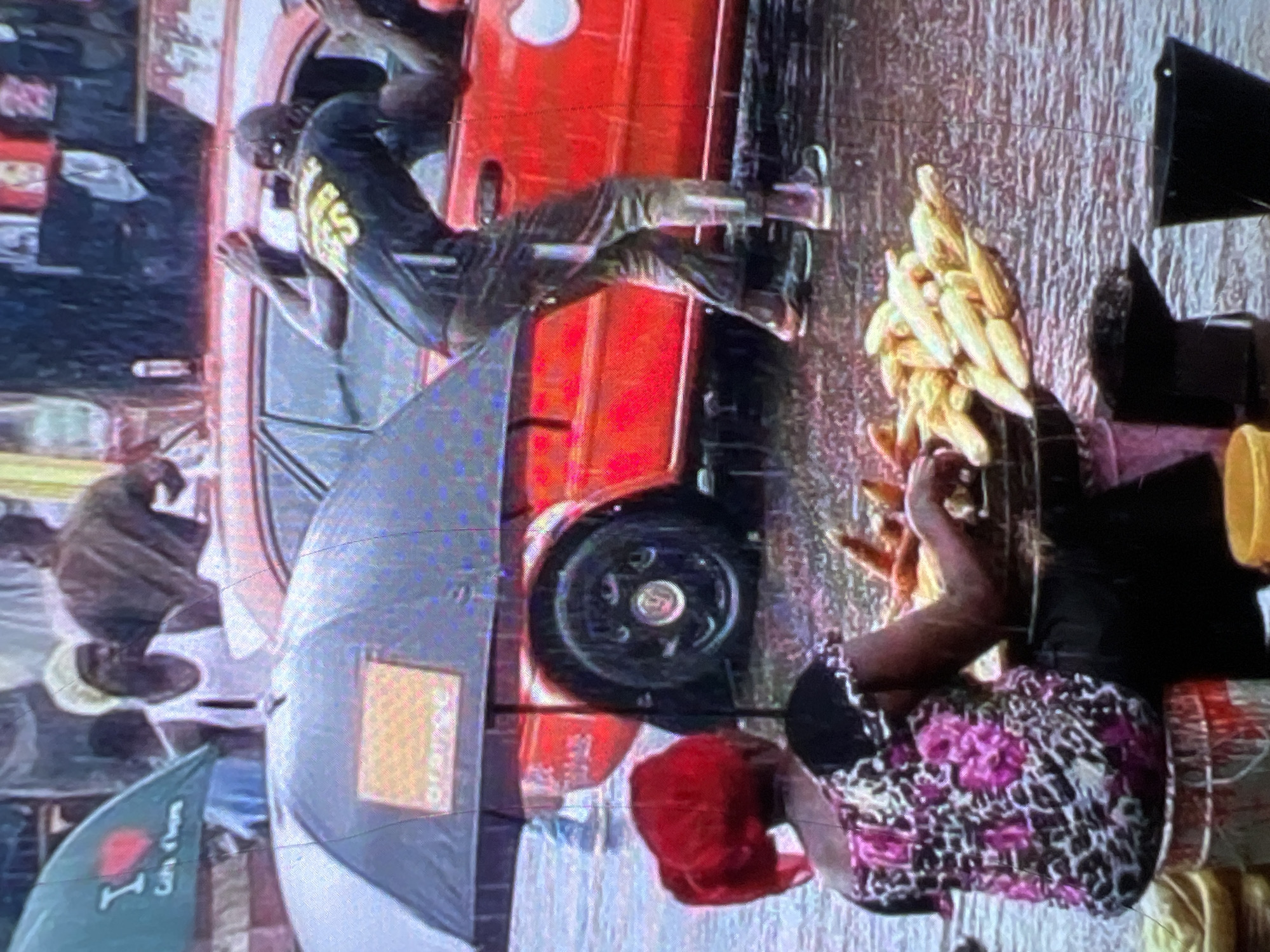 The image depicts a street scene in Lagos, Nigeria, during a downpour. A woman wearing a red headscarf and a patterned top is seated, facing away from the camera, attending to a pile of corn cobs. She is positioned near a stall or cart, indicated by the basket and buckets around her. In the background, a red car is visible, with its door open. A person, wearing a dark t-shirt with yellow lettering, leans out of the car window. An umbrella, with an "Orange" logo, is open, partially obscuring the car and the woman. Other individuals are visible in the background, some under umbrellas or in what appear to be makeshift shelters. The ground is wet, reflecting the rainy conditions. The overall atmosphere is busy and likely reflects the resilience of street vendors and daily life in Lagos, even amidst inclement weather.