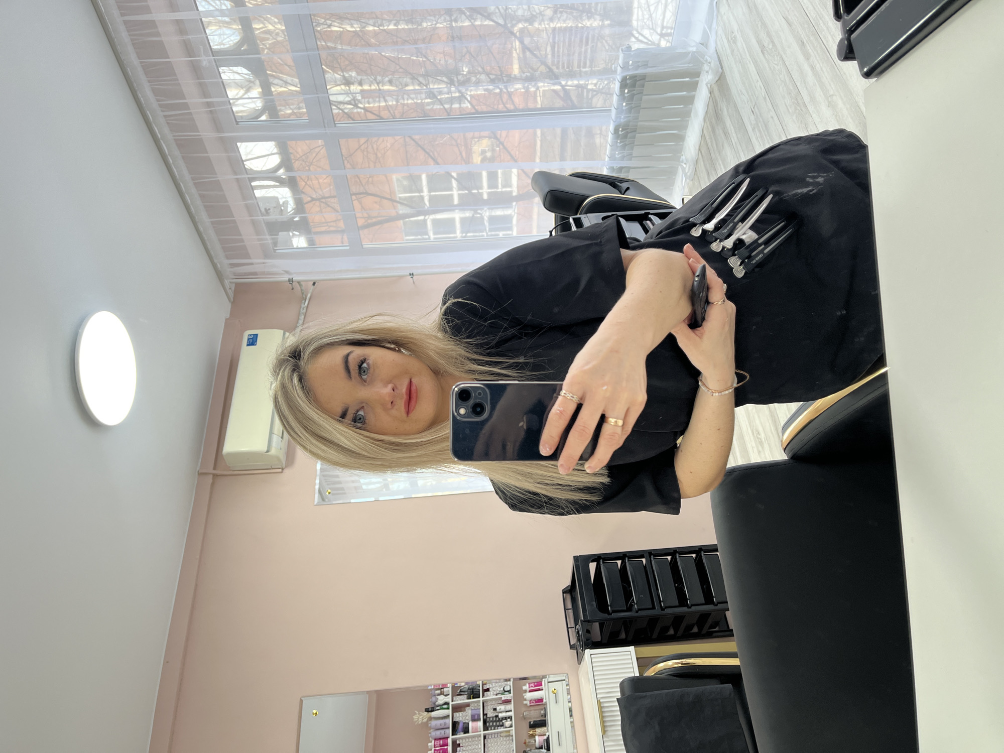 A blonde female, estimated to be in her 20s or 30s, is captured in a mirror selfie within a hair salon located in Chaikovsky, Russia. She wears a black short-sleeved uniform or tunic and a black apron adorned with multiple silver hair clips on the lower right side. Her right hand holds a dark-colored smartphone, positioned to take the photograph, while her left arm rests across her body. Rings are visible on her fingers and bracelets on her left wrist. She looks directly towards the camera with a neutral expression.
The salon interior features light pink walls and a white ceiling with a circular flush-mount LED light fixture. Light-colored wooden or laminate flooring is present. To the subject's left, a white vanity with ridged drawer fronts and gold handles holds a small white vase with decorative items; a reflection in a mirror shows shelves stocked with numerous hair care products in various packaging. A black rolling salon cart with multiple drawers is positioned between the vanity and the subject. Black styling chairs with gold accents are visible, with one directly behind the subject. A white, wall-mounted air conditioning unit is fixed high on the right wall.
On the right side of the image, a large window with white frames is covered by sheer white curtains. Through the window, leafless trees and a multi-story reddish-brown brick building with several windows are visible, suggesting an exterior urban environment during a cooler season. A white radiator is installed on the wall directly beneath the window. The foreground shows a partial reflection of a white countertop or table surface.