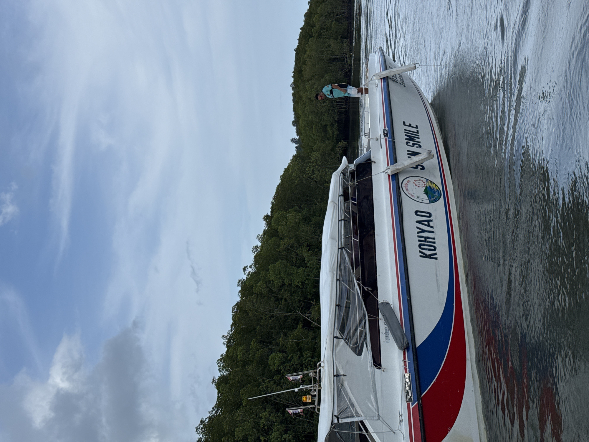 A speed boat named "S'N SMILE" is docked on the water near a tree-lined shore. The boat is white with red and blue accents and has a canopy. A man in a blue shirt and white shorts stands on the back of the boat looking down. The water is calm and reflects the sky and the boat. The sky is cloudy and the trees are dense and green. The location is Pa Khlok, Thailand.
