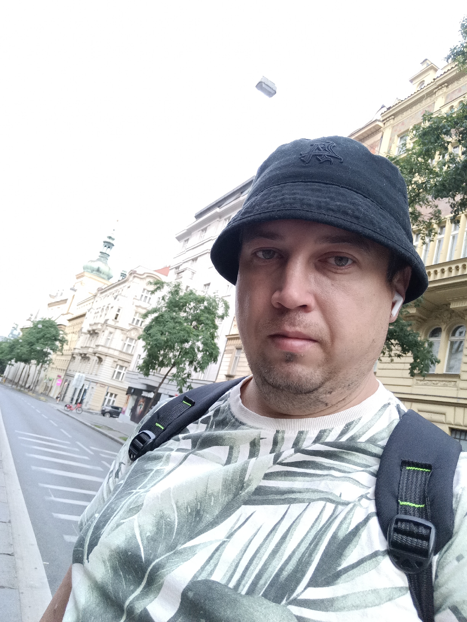 An outdoor, eye-level, medium selfie shows a man in his 30s wearing a black bucket hat with an embroidered logo, a white t-shirt with a green palm leaf pattern, and a black backpack with green accents. He has light skin, brown hair, and is looking directly at the camera. He is wearing white wireless earbuds in his right ear. The man is standing on a sidewalk with a street and buildings behind him. The buildings are European-style architecture with ornate details and a dome-shaped tower. The street is paved with a crosswalk visible in the foreground. There is a red bicycle parked on the side of the street and a black car in the distance. The sky is overcast. The scene appears to be in Prague, Czechia.