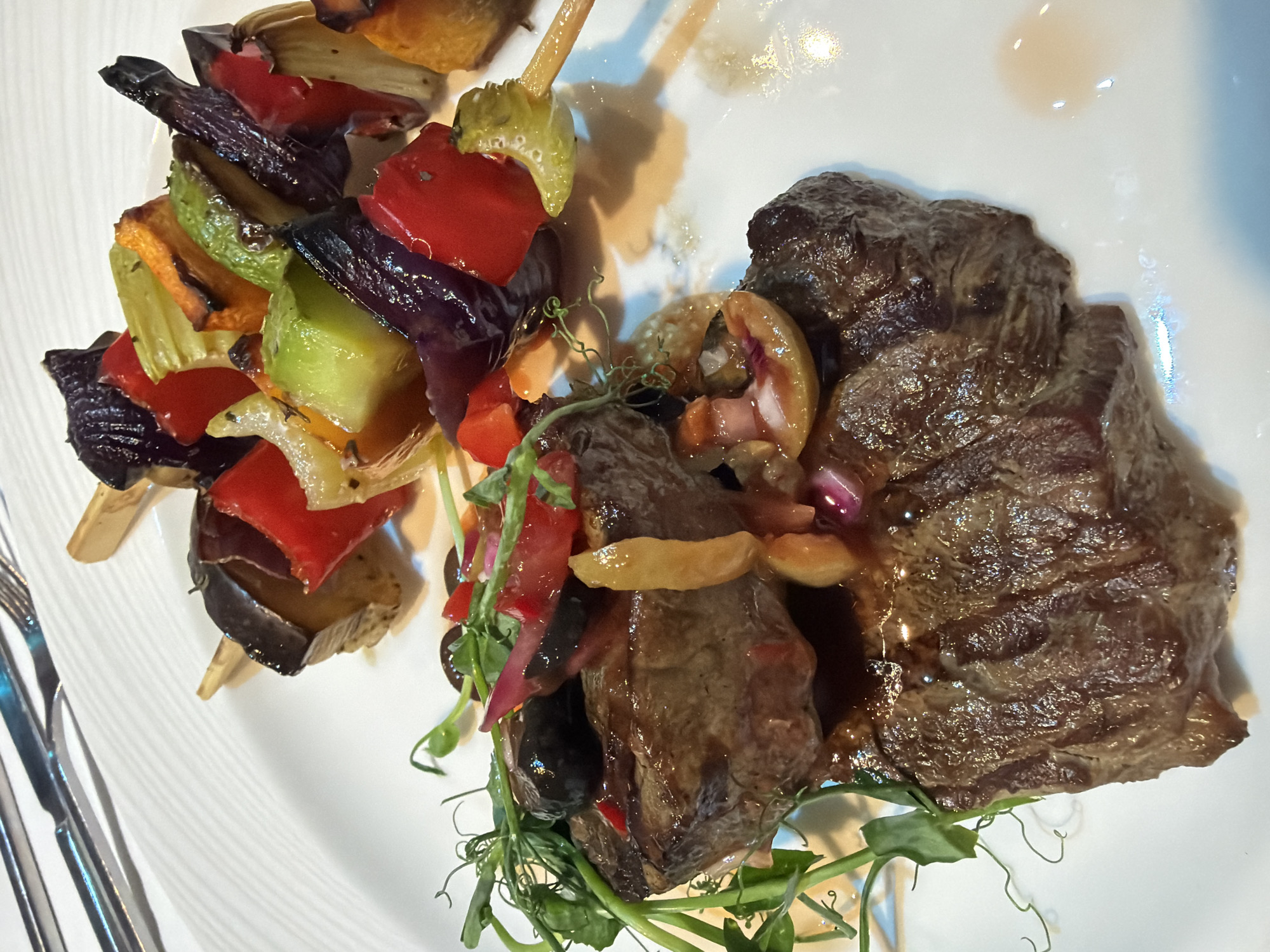 This media file presents an overhead, close-up view of a meticulously plated gourmet meal. The central subjects are two pieces of richly colored, cooked meat, likely beef steak, which appear seared and glazed with a dark, savory sauce. The larger piece of meat exhibits a textured, cooked surface. The second, more irregularly shaped piece, is adorned with various toppings, including diced red bell peppers or tomatoes, sliced golden-brown mushrooms or onions, and slivers of red onion. Lush green microgreens, possibly pea shoots, delicately encircle the meat, adding a fresh contrast.
To the left of the meat are two wooden skewers showcasing vibrant, grilled or roasted vegetables. These skewers feature substantial chunks of red bell pepper, green zucchini, purple eggplant, and possibly yellow bell pepper, all bearing subtle char marks indicative of cooking.
The meal is served on a pristine white ceramic plate, which features a faint, linear embossed pattern. A silver fork and knife are partially visible in the bottom-left corner, resting alongside the plate. The overall presentation is refined, suggesting a dining experience in an indoor setting, located in Moscow, Russia.
