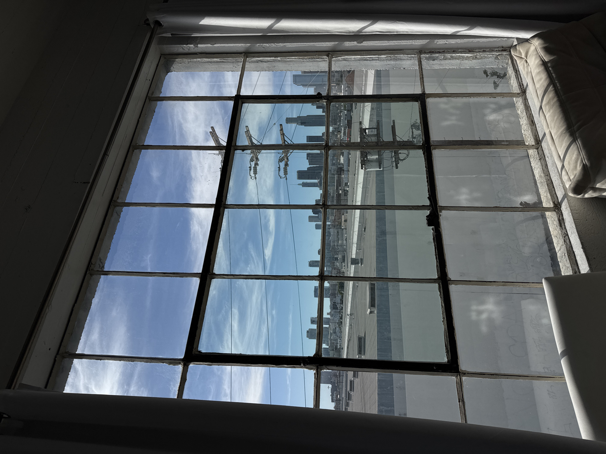 The image depicts a view of the Los Angeles skyline through a gridded window, likely from an interior space. The skyline includes several high-rise buildings, with construction cranes visible among them. The sky is blue with scattered clouds. The window itself is old, with a white frame and multiple panes. Some of the glass panes appear dirty or textured. Part of the interior of the space is visible in the foreground, including a white object, possibly furniture. The image is slightly angled.