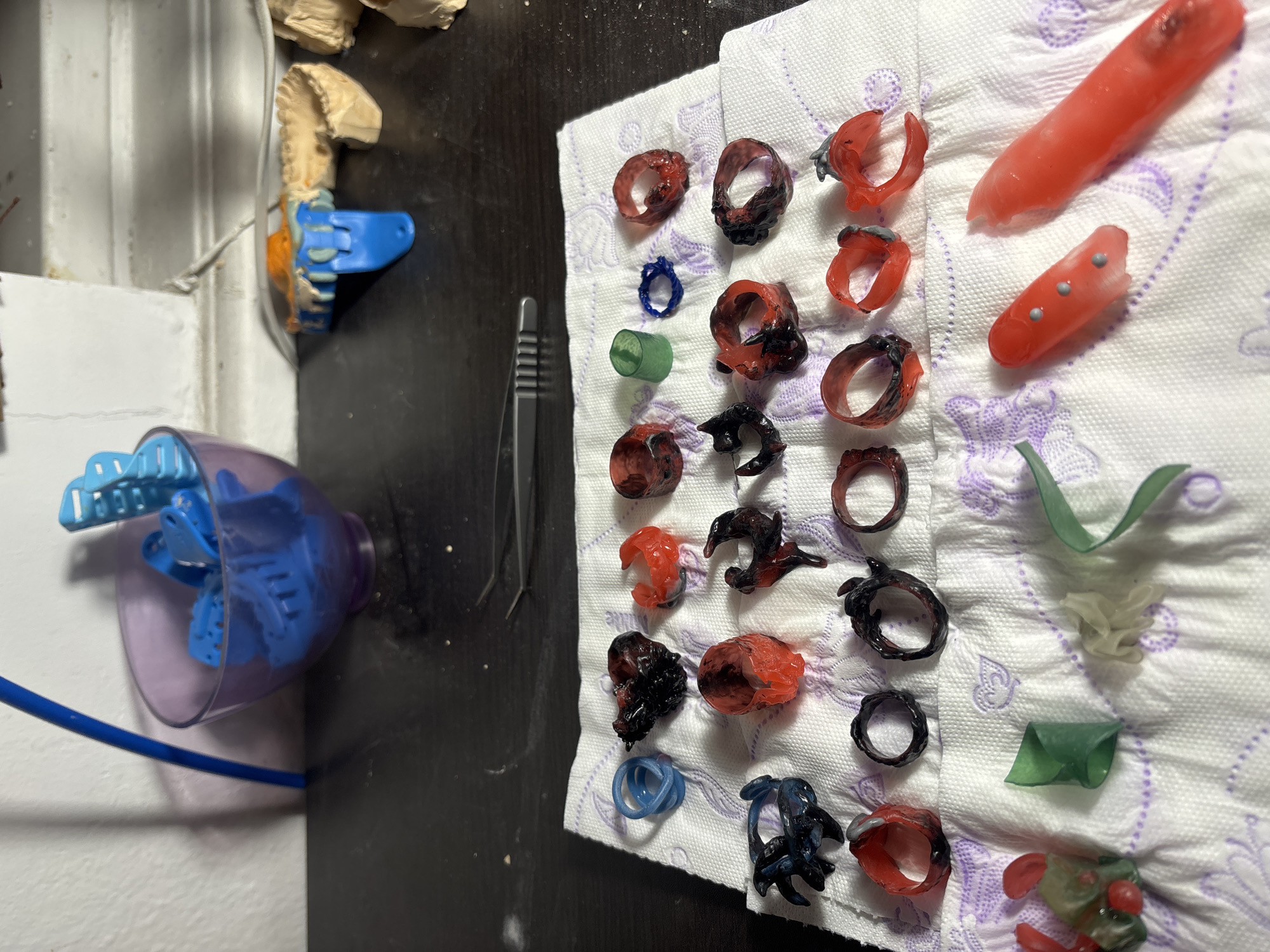 The image depicts a tabletop scene. A white napkin with a lavender pattern sits to the right, holding an assortment of castable wax prototypes for jewelry, including rings, organic shapes, and cylindrical forms in shades of red, black, blue, green and clear. To the left of the napkin lies a black surface. An open clear and purple plastic basin containing blue plastic clips sits on the edge of the black table, a blue hose trailing from it. A metal tool rests on the black table near the napkin. A plaster dental mold with a blue clasp is in the upper left corner of the image, resting near the wall. The environment suggests a jewelry making or dental prototyping workspace. The location is indeterminate; there is no data suggesting that the picture was taken in Sofia, Bulgaria. There are no people present in the frame.