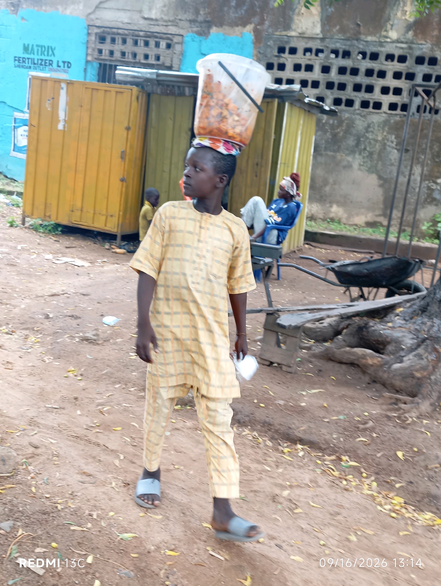A young boy walks on a dirt path in Shendam, Nigeria, carrying a large bucket of food on his head. He wears a yellow and brown checkered traditional outfit and grey sandals. In the background, a yellow shipping container is visible, along with a building with a barred window. A person sits in a blue chair behind the boy, and a wheelbarrow is present in the mid-ground. The ground is covered in dirt and scattered leaves. A timestamp in the lower right corner reads "09/16/2026 13:14", and "REDMI 13C" is in the lower left corner.