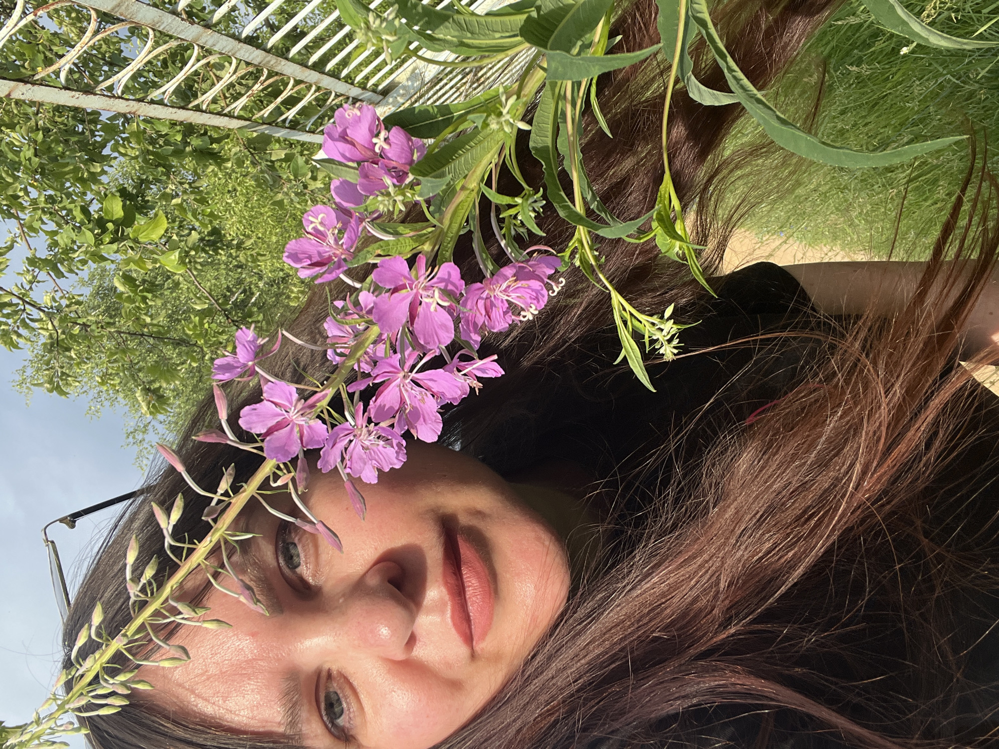 The media file shows a close-up, rotated shot of a person's face, with purple flowers and greenery in the foreground. The person has long, brown hair and is wearing glasses. Their eyes are open, and they appear to be looking towards the camera with a gentle smile. The lighting suggests it is daytime, possibly late afternoon due to the warm tones. The background includes lush green foliage and a white, decorative metal fence, hinting at an outdoor setting such as a garden or park. The overall mood is relaxed and natural, with the flowers adding a touch of delicate beauty to the portrait. The location context is Noviy, Russia. No visible text is present in the media file.