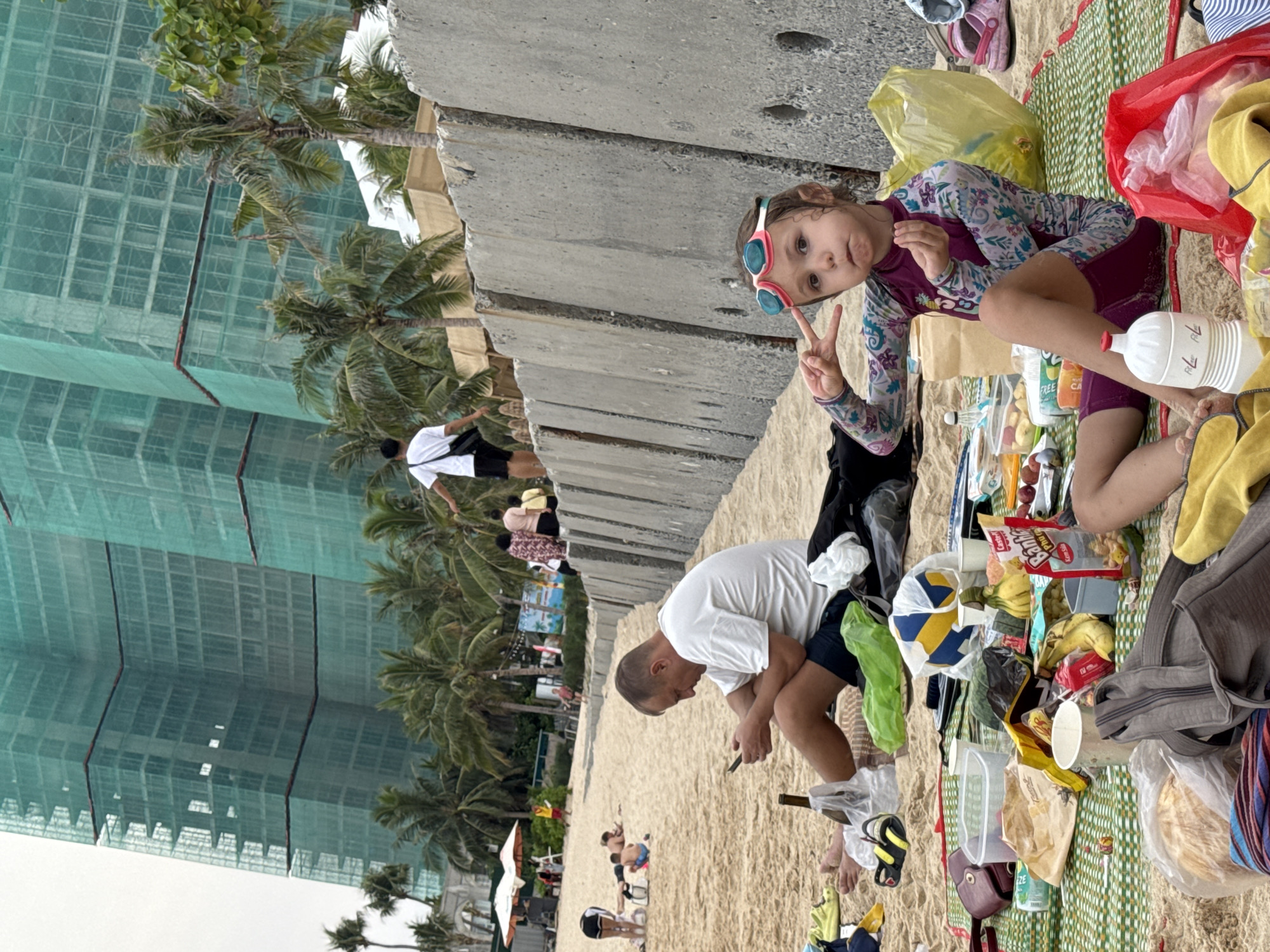 The image shows a beach scene at Bai Nha Trang Ward, Vietnam, as evidenced by the construction and palm trees in the background. A young girl wearing goggles sits cross-legged on a green and white checkered mat, giving a peace sign with her right hand. Various food items, containers, and bags surround her on the mat. An adult male in a white t-shirt and dark shorts sits nearby, looking at a mobile phone. Other people can be seen in the background along the beach. A concrete wall runs along the edge of the beach, separating it from the buildings.