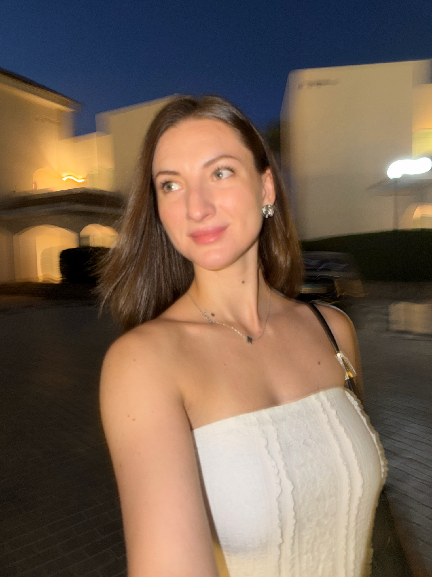 A young woman with shoulder-length brown hair and light skin is pictured outdoors at dusk. She is wearing a white strapless top and a silver necklace with a small pendant. She is also wearing a sparkly earring on her left ear. Her skin has a few freckles. The background consists of buildings with warm lighting, suggesting an urban or resort setting at night. The image quality is slightly blurred due to motion, but the woman's face is clearly visible as she looks to her left with a slight smile. The location is identified as Sharm El Sheikh, Egypt.